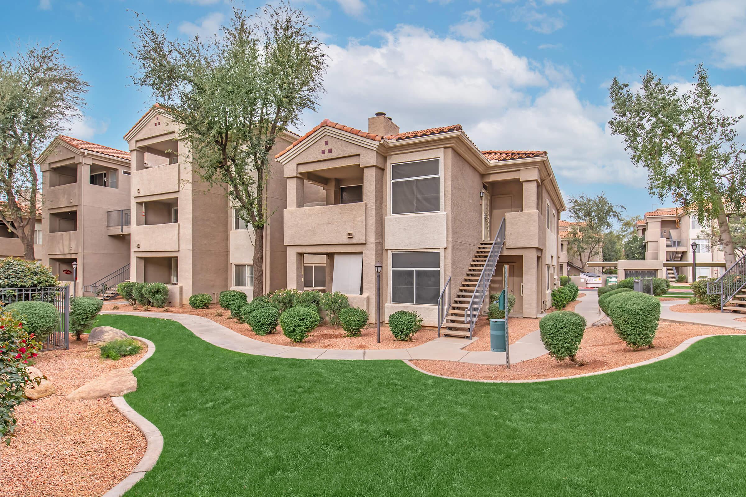 A well-maintained apartment complex featuring tan-colored buildings with tile roofs, surrounded by lush green grass and neatly trimmed shrubs. The layout includes walking paths and small trees, creating a welcoming outdoor space. Blue sky with fluffy clouds overhead.