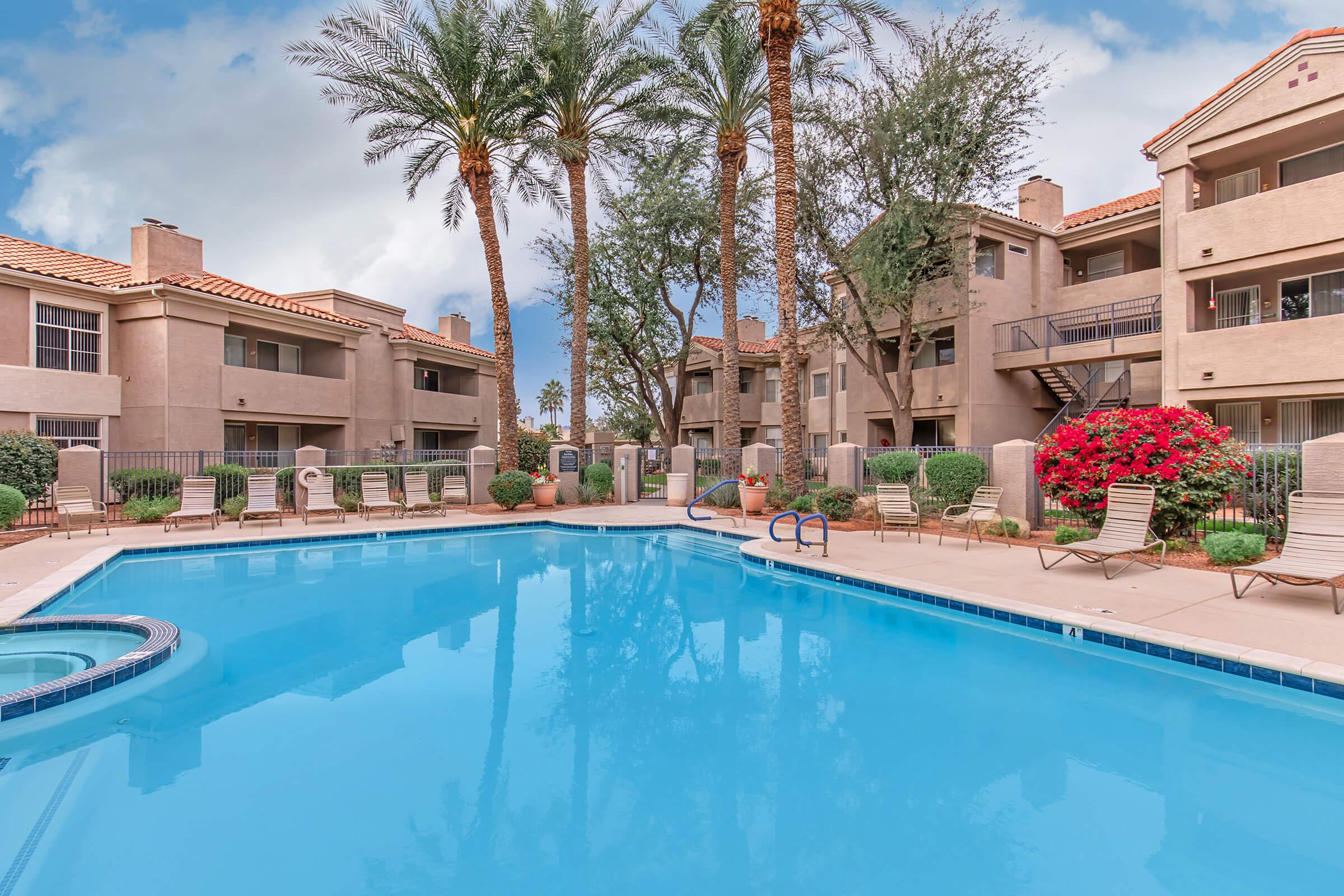 A serene pool area surrounded by palm trees and vibrant pink flowers. The pool features a jacuzzi and is framed by beige apartment buildings with balconies. Sun loungers line the poolside, creating a relaxing atmosphere perfect for leisure and enjoyment.