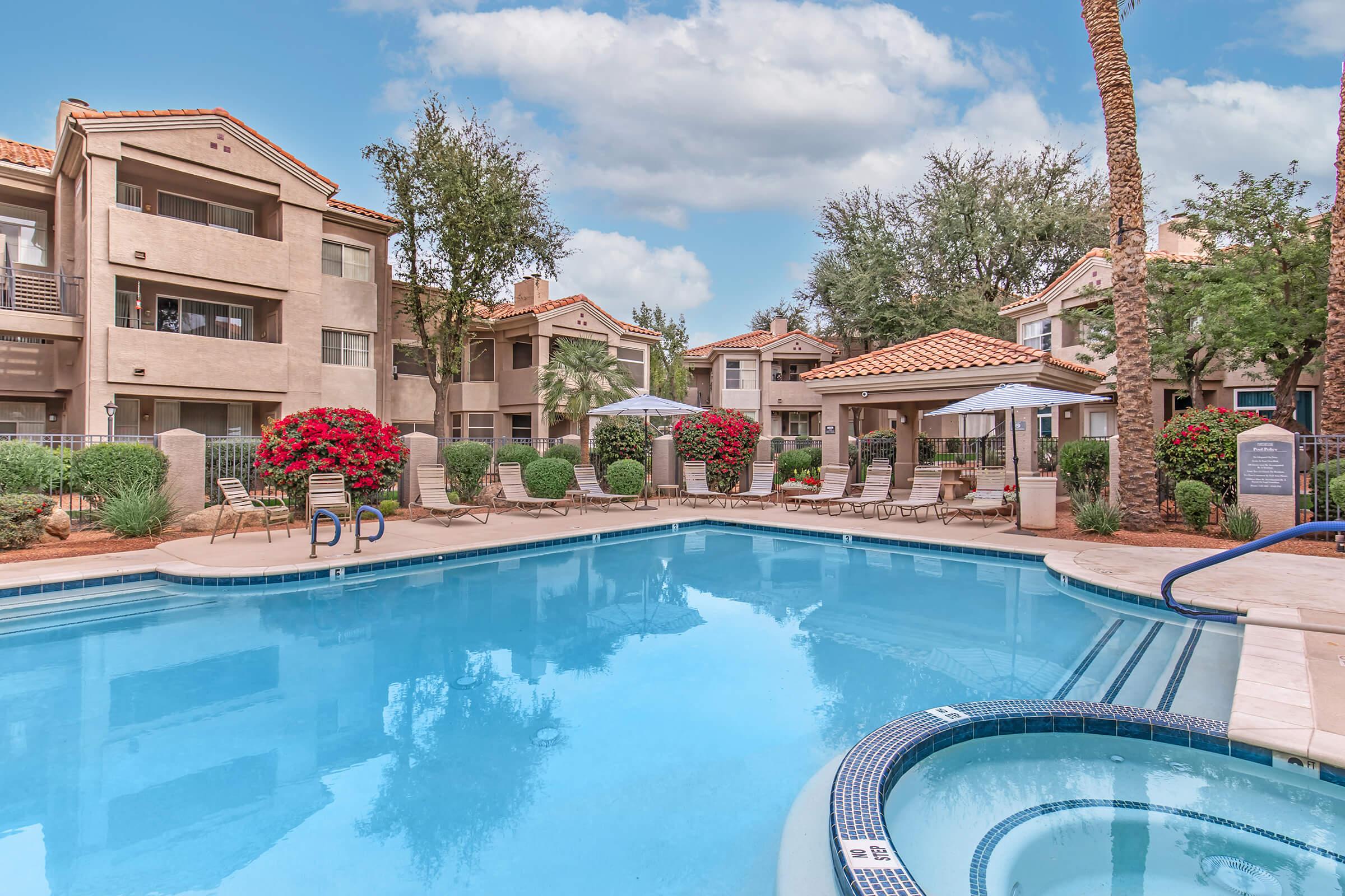 A serene apartment complex featuring a clear blue swimming pool surrounded by lounge chairs and vibrant flowering shrubs. The buildings, adorned with terracotta roofs, are set against a backdrop of palm trees and blue skies, creating a peaceful outdoor retreat.