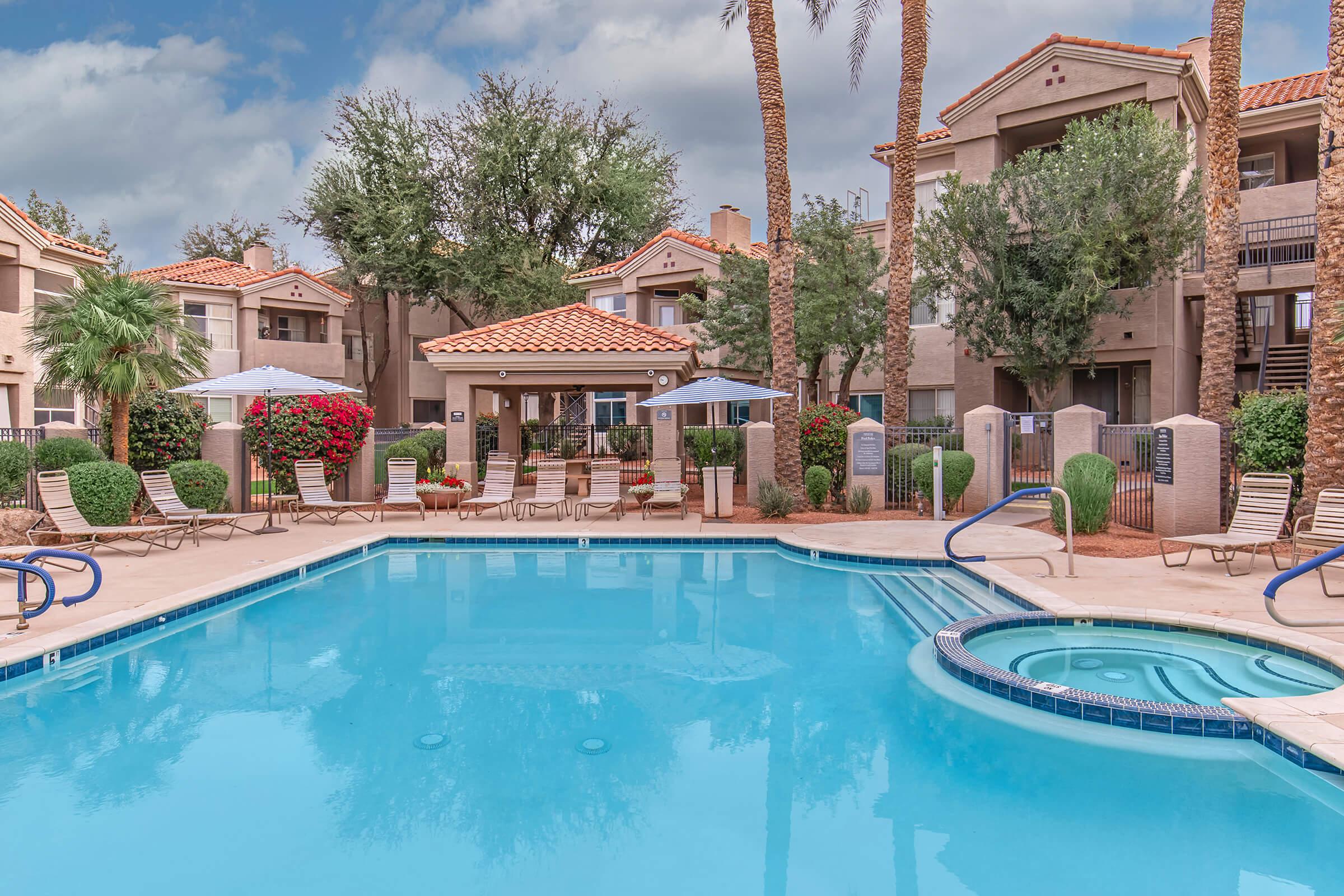 A serene pool area surrounded by palm trees and blooming bushes. Lounge chairs are arranged around the pool, and umbrellas provide shade. In the background, multi-story apartment buildings feature terracotta roofs. The sky is partly cloudy, creating a tranquil atmosphere.