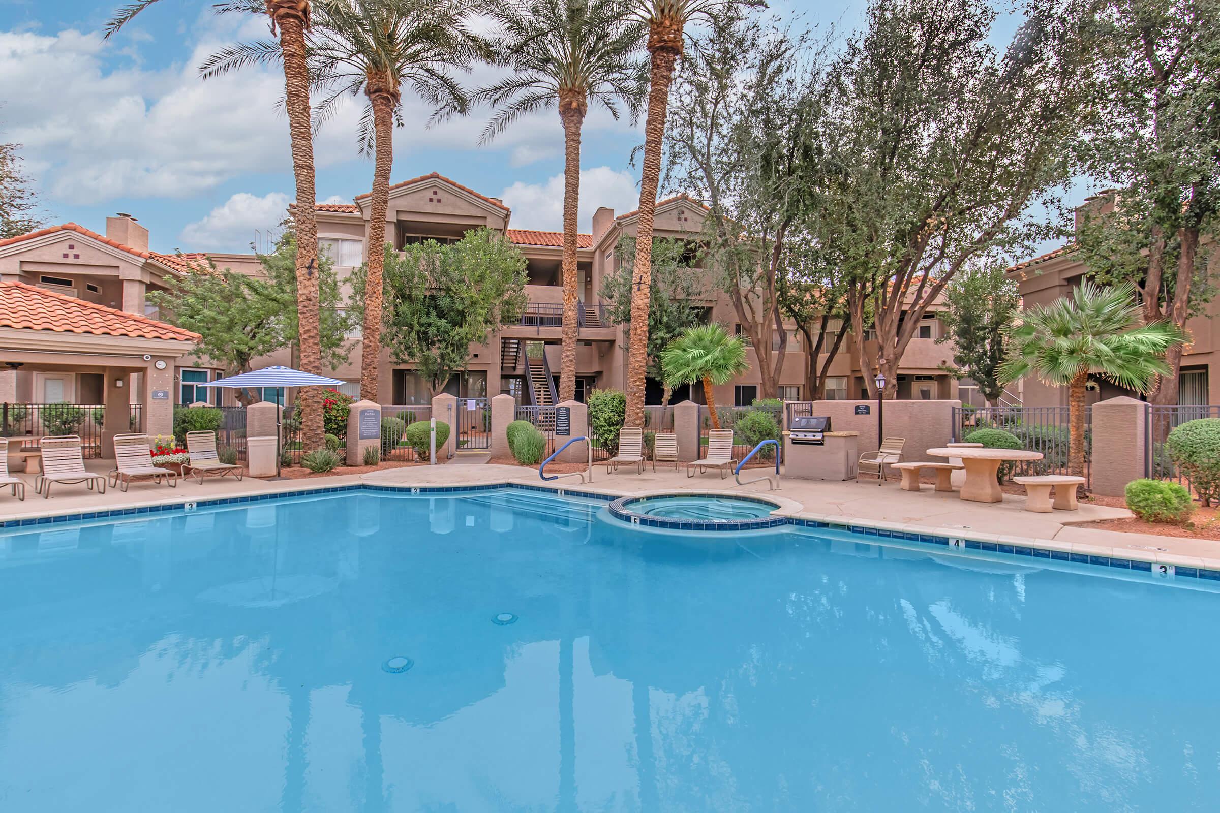 A clear blue swimming pool with a spa area surrounded by palm trees. In the background, there are beige apartment buildings with balconies and lush green landscaping. Several lounge chairs are arranged around the pool, along with umbrellas for shade, creating a relaxing outdoor atmosphere.