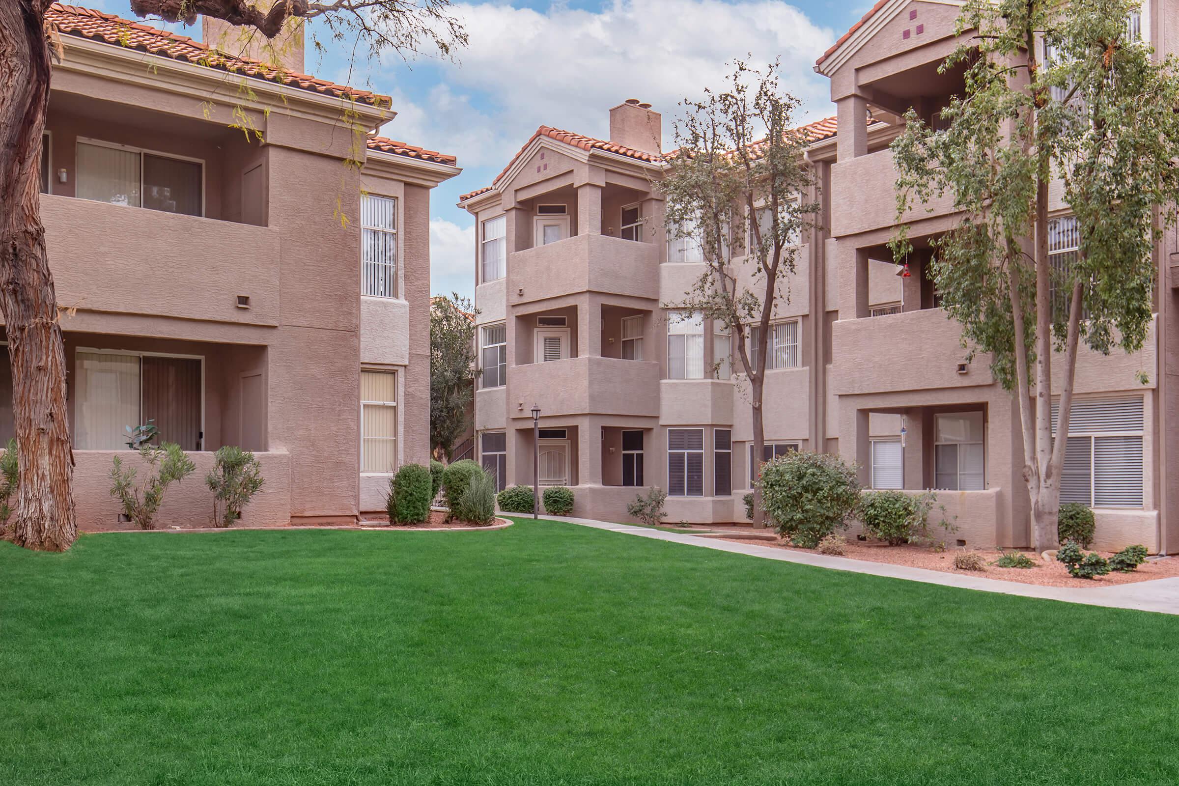 View of a residential area featuring two beige apartment buildings with balconies, surrounded by a well-maintained green lawn and small shrubs. The sky is partly cloudy, suggesting a pleasant day. The layout includes a walkway between the buildings leading to the lawn.