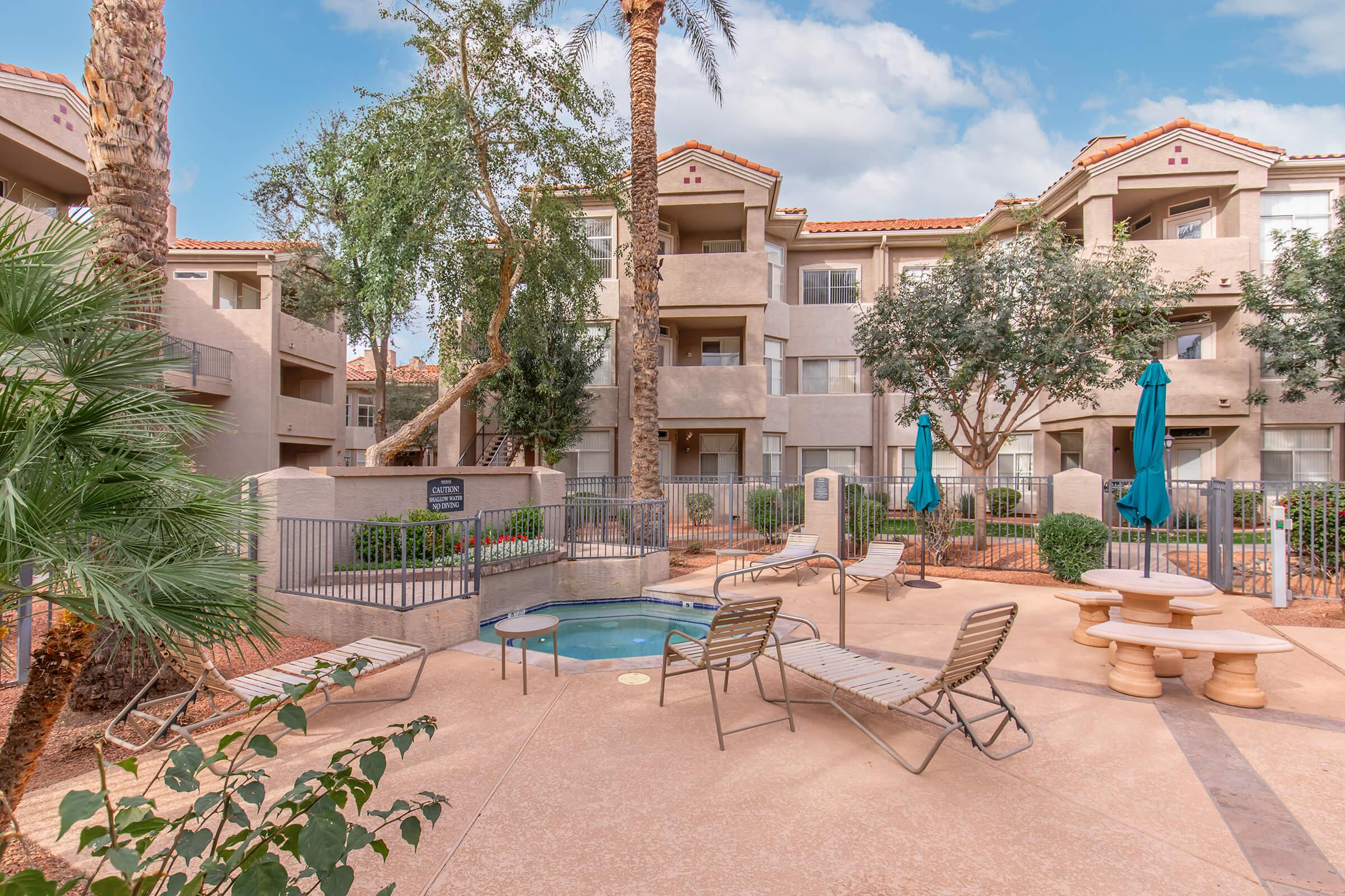 A serene apartment courtyard featuring a small hot tub, lounge chairs, and shaded tables. Lush greenery, palm trees, and bright blue umbrellas create a relaxing outdoor space. The backdrop includes multiple residential buildings under a partly cloudy sky.