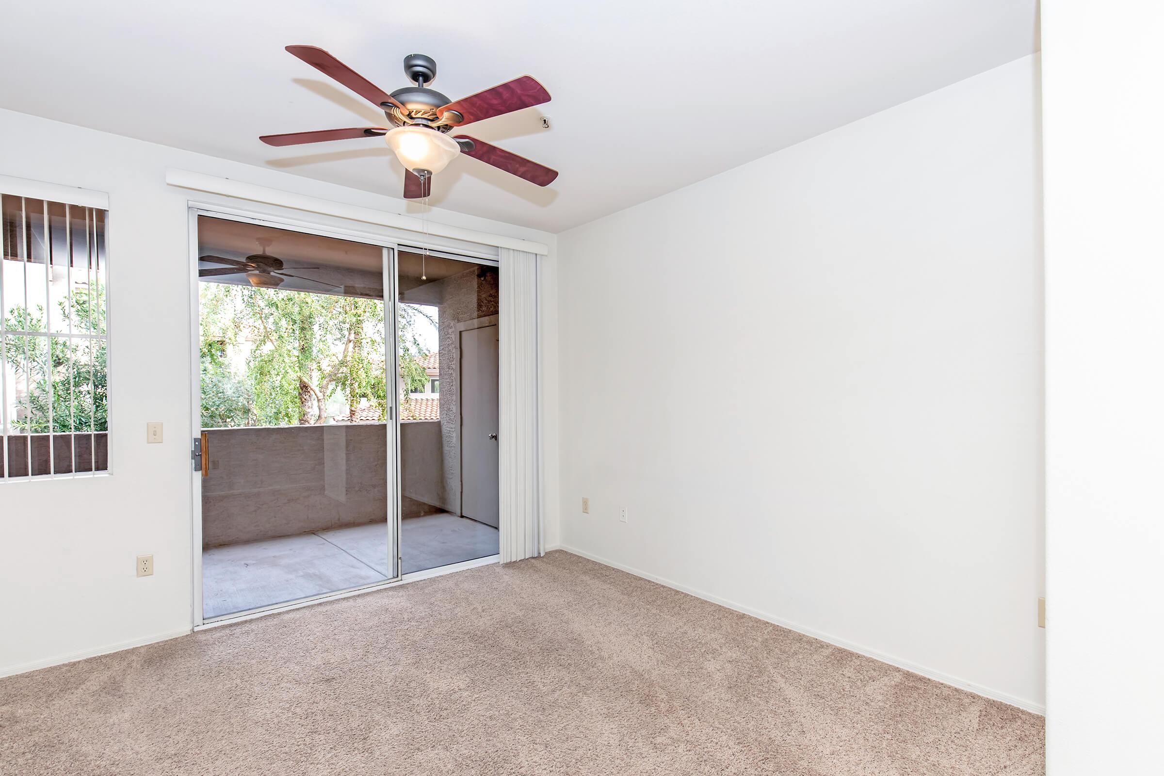 A spacious, empty room with beige carpet and white walls. A ceiling fan with wooden blades hangs above. Large sliding glass doors lead to a balcony, allowing natural light to enter. The room has a minimalist design, suitable for various furnishings and décor.