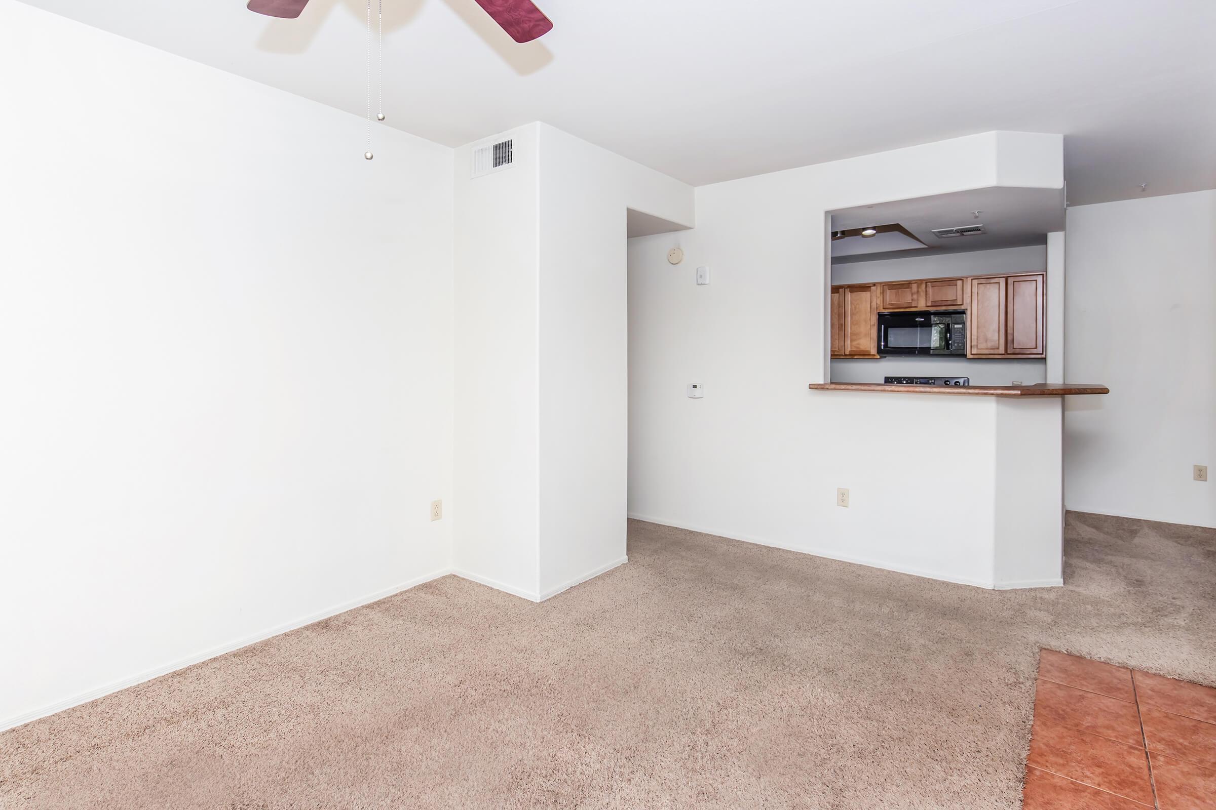 Empty living room area with light-colored walls and carpet. A ceiling fan is visible, and there is a partial wall leading to a small kitchen with wooden cabinets and modern appliances. The space appears bright and open, suitable for furniture arrangement.