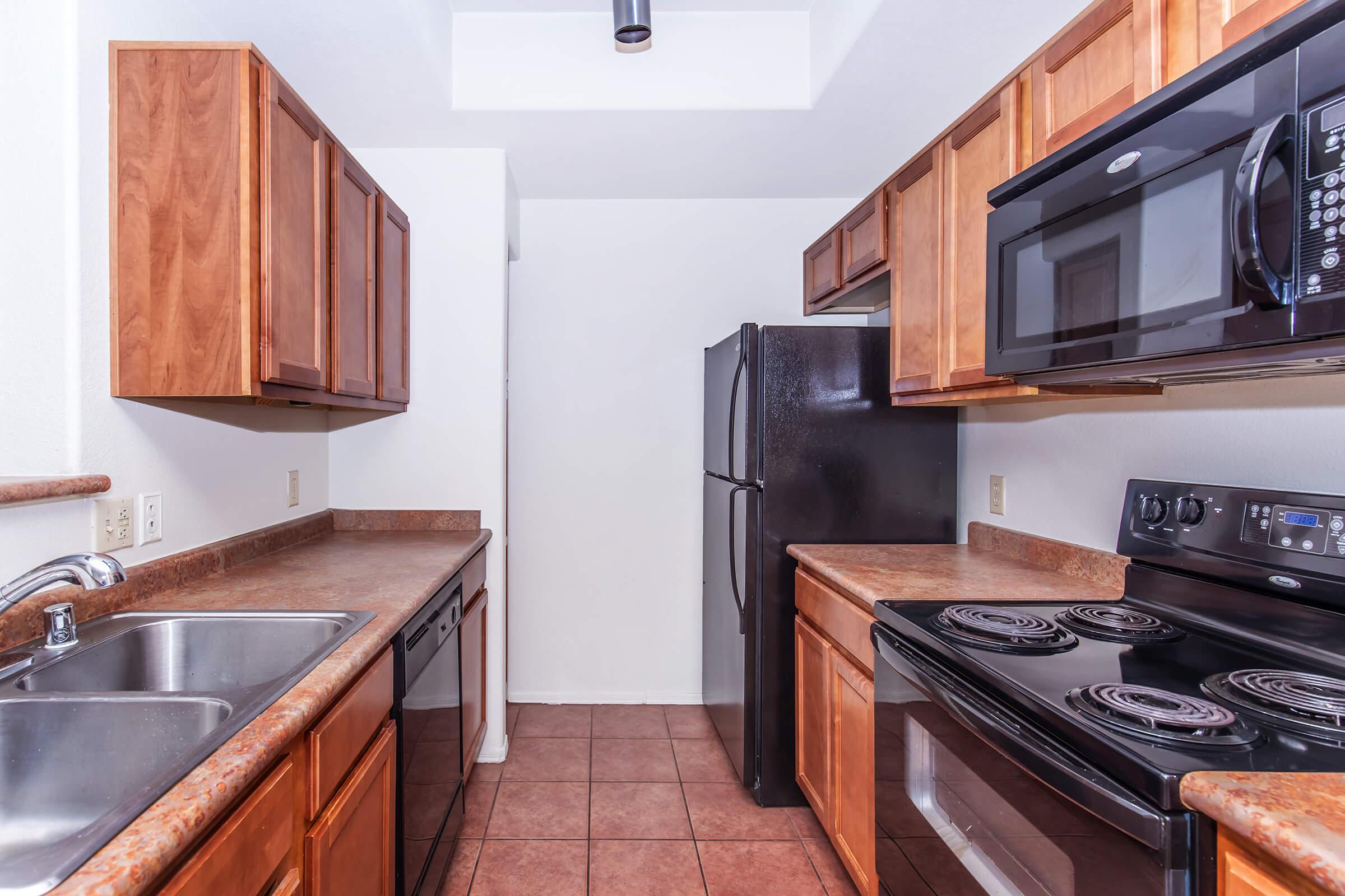 A modern kitchen featuring wooden cabinetry, a double sink, a black refrigerator, an electric stove, and a microwave. The countertops have a brown finish, and the floor is tiled. The overall color scheme is warm and inviting, highlighting a cozy yet functional cooking space.