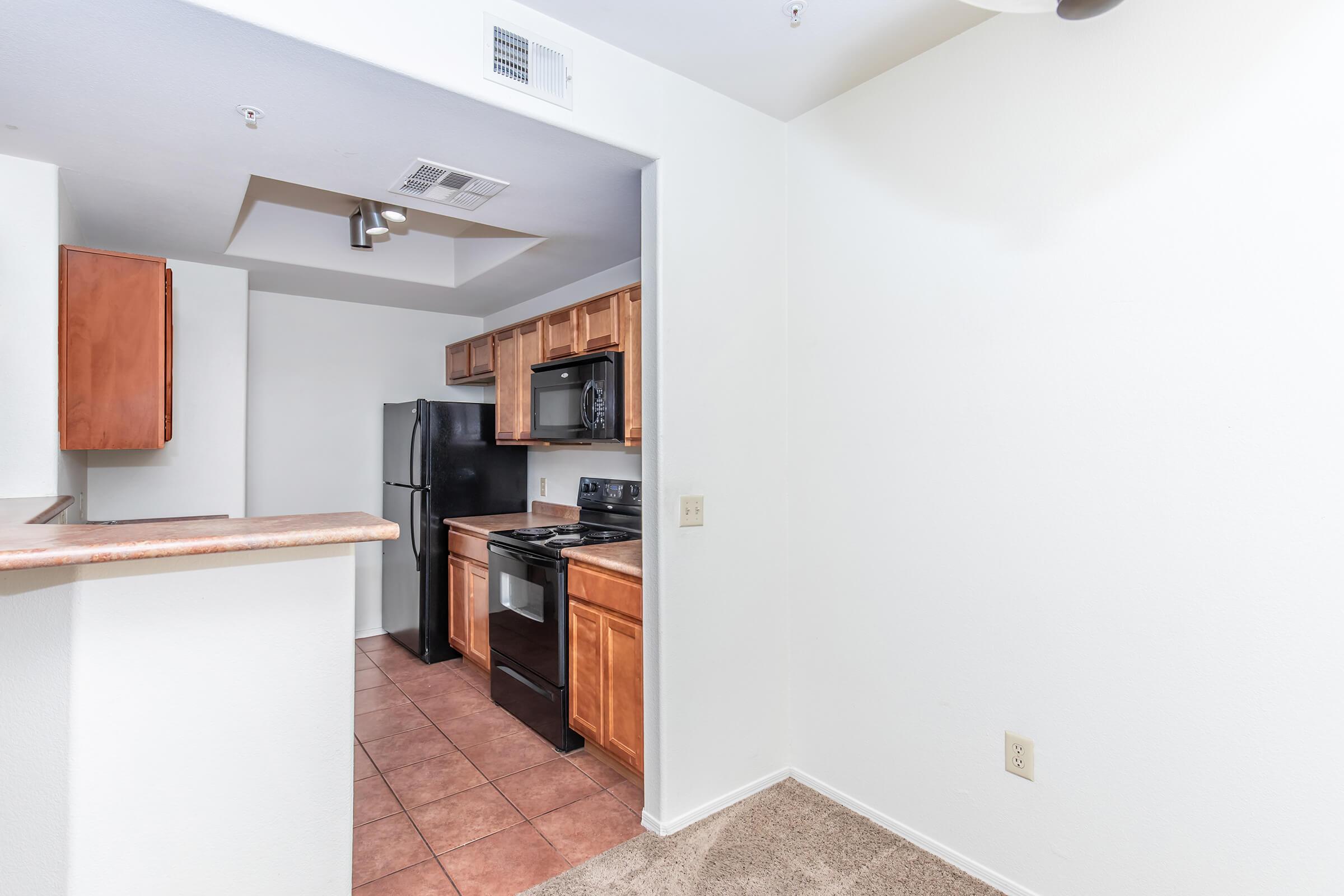 Modern apartment kitchen featuring wooden cabinets, black appliances including a refrigerator and stove, and a countertop dividing the kitchen from the living area. The flooring is tiled, and the walls are painted white, creating a clean and spacious atmosphere.