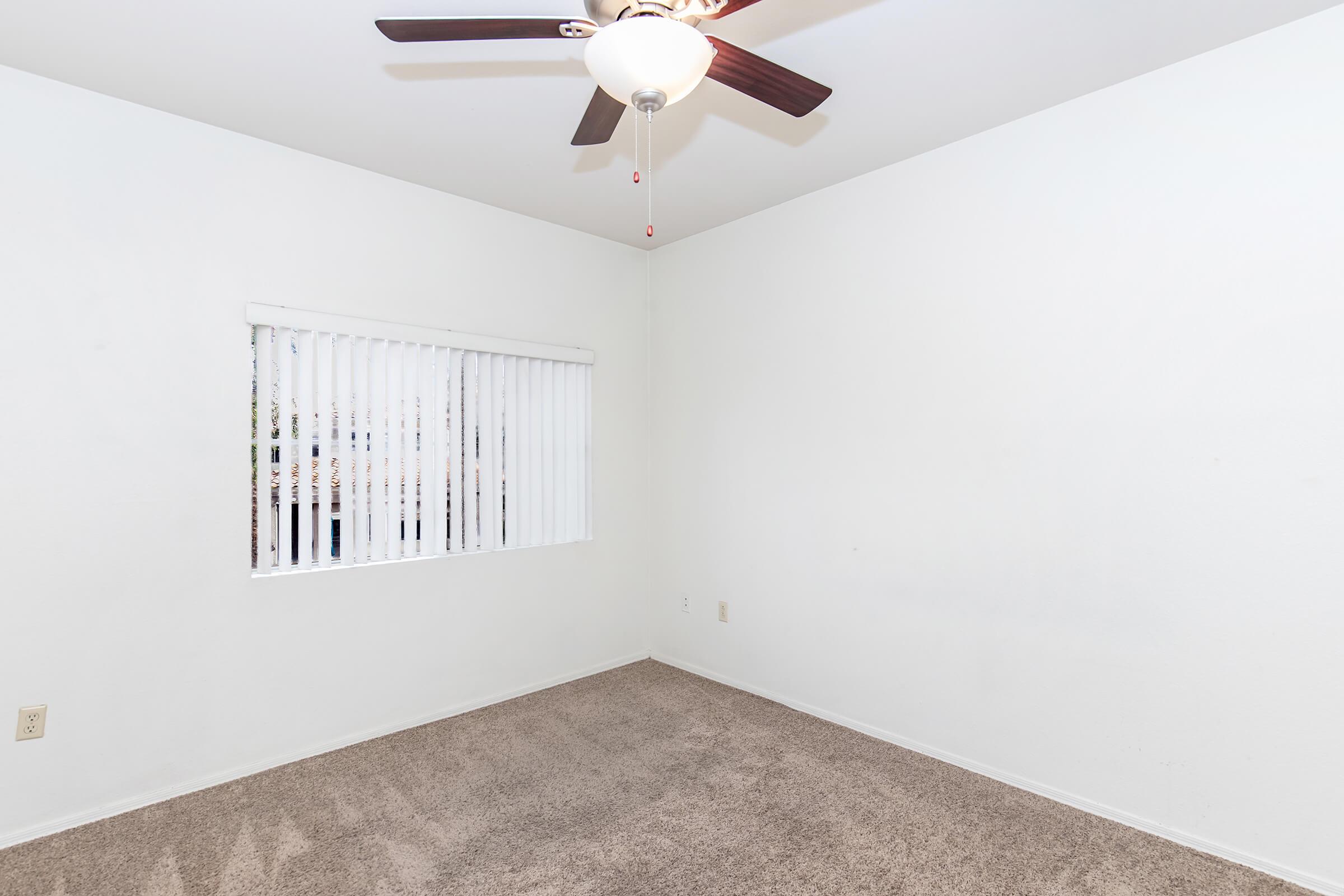 A sparsely furnished room with beige carpet and white walls. There is a ceiling fan with a light fixture and a window covered by white blinds, allowing natural light to enter the space. The room appears clean and empty, suggesting it might be used for various purposes.