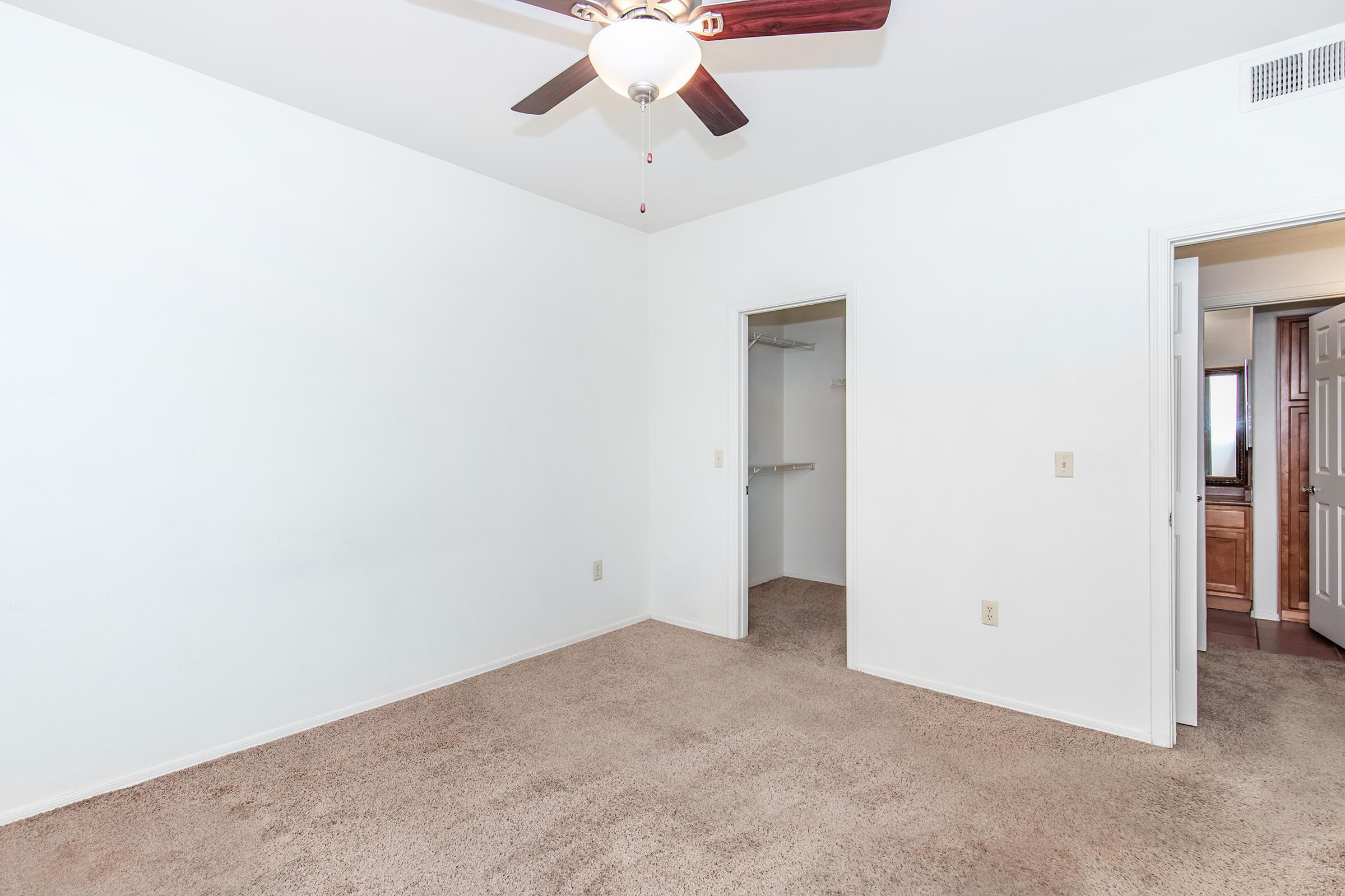 Empty room with light beige carpet, white walls, and a ceiling fan. There is a closet opening on the left and a door leading to another room on the right. The space is well-lit and unfurnished, creating a blank canvas for interior design.