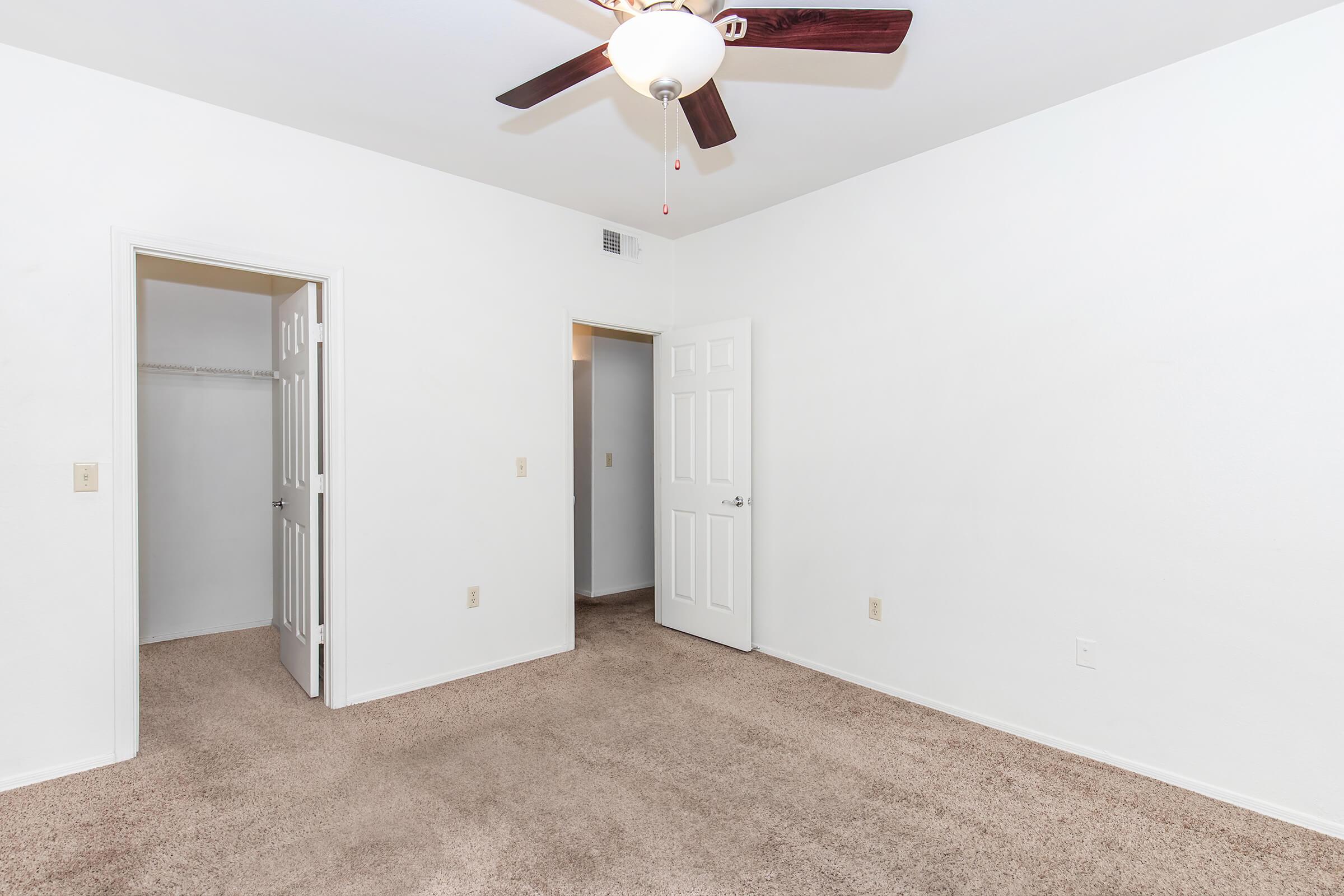 A sparse room featuring light-colored walls and a ceiling fan. There are two doors – one leading to a closet and the other to what appears to be another room. The floor is covered with light beige carpet, giving a neutral and open feel to the space.