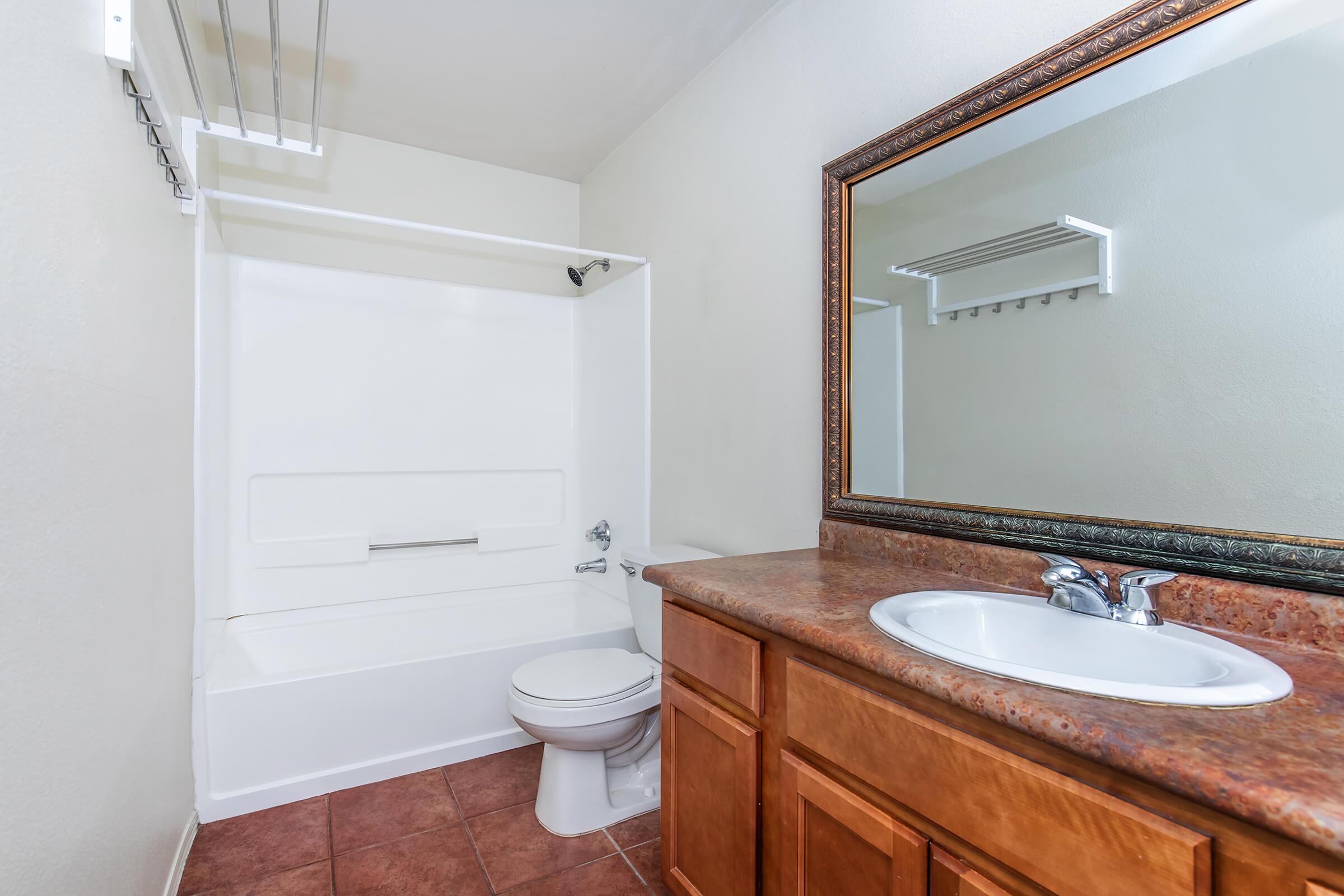 A clean bathroom featuring a white bathtub and shower combination, a toilet, and a wooden vanity with a sink. There is a large mirror above the sink and a towel rack on the wall. The flooring is tiled in a warm color, and the walls are painted in a soft, neutral shade.
