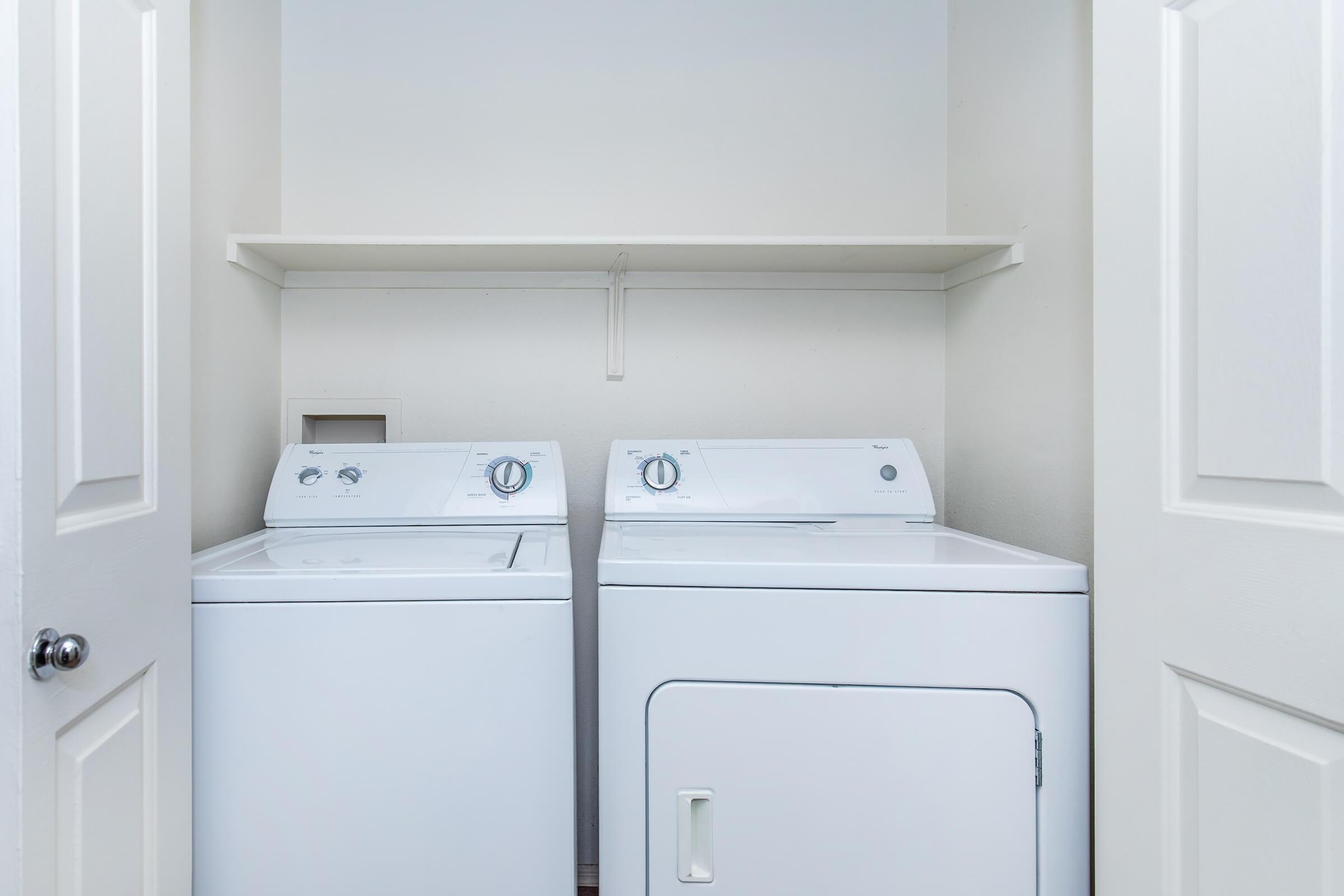 A laundry room featuring a white washing machine and dryer side by side, with a shelf above for storage. The room is well-lit and has light-colored walls, creating a clean and simple appearance. The doors to the laundry space are partially open.
