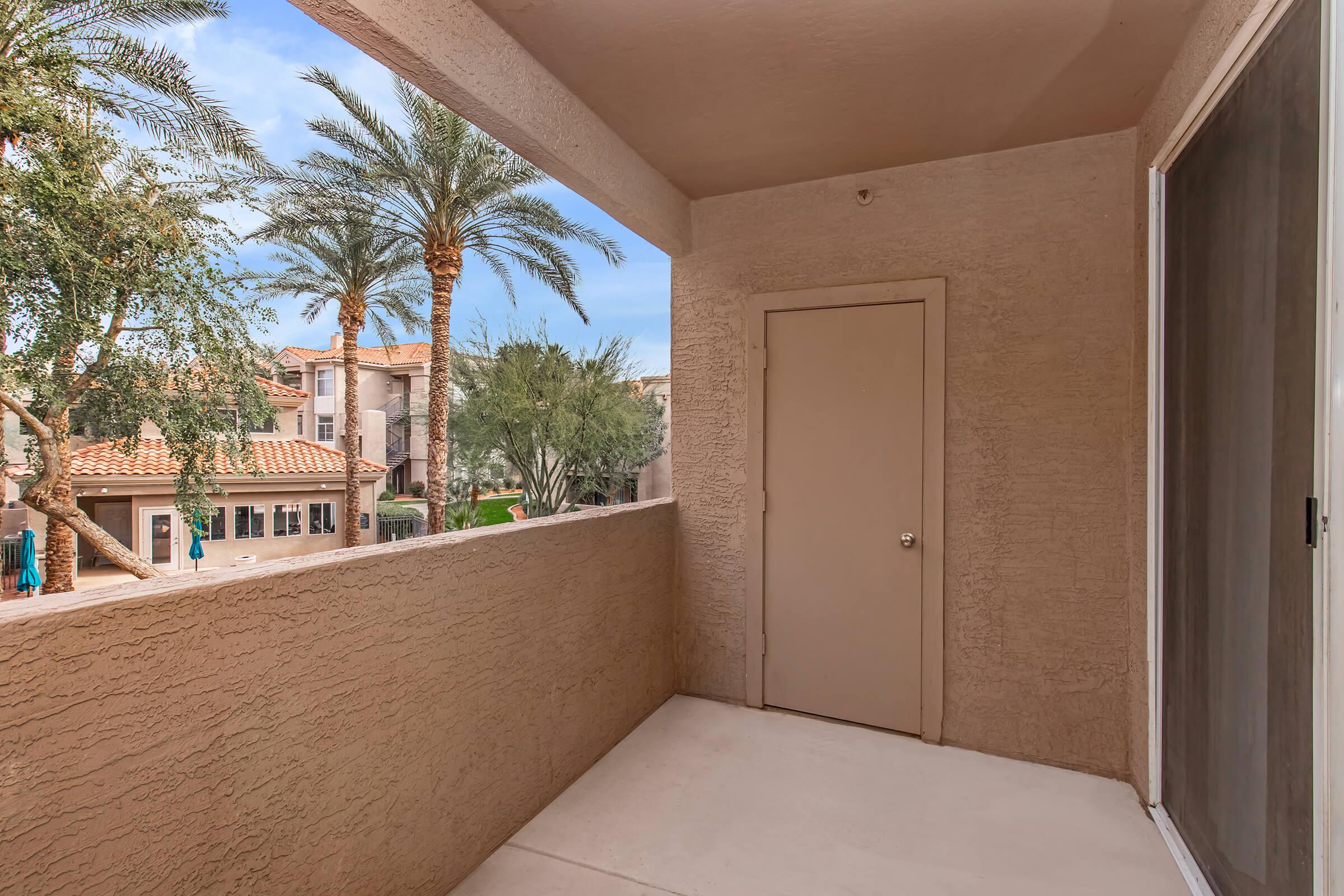 A view from a balcony featuring a beige wall, a closed door, and palm trees outside. The scene showcases a sunny day with clear skies and nearby buildings visible in the background, creating a serene outdoor atmosphere.