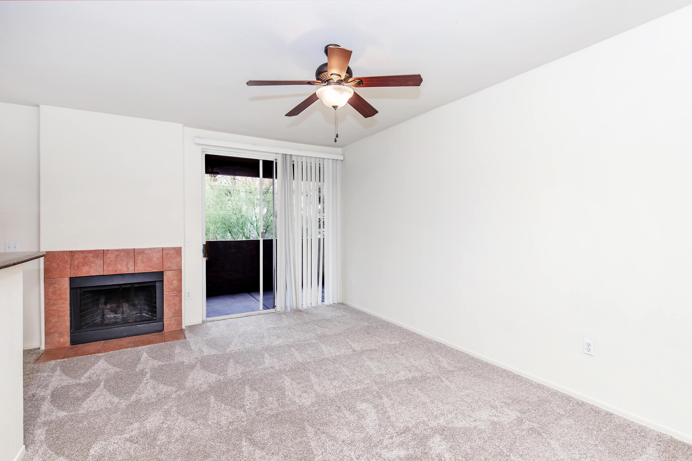 Spacious living room featuring beige walls, a ceiling fan with wooden blades, and a fireplace with a tiled facade. Large sliding glass door leads to a balcony, providing natural light. The room has plush, light-colored carpet and a cozy atmosphere, perfect for relaxation and entertaining.