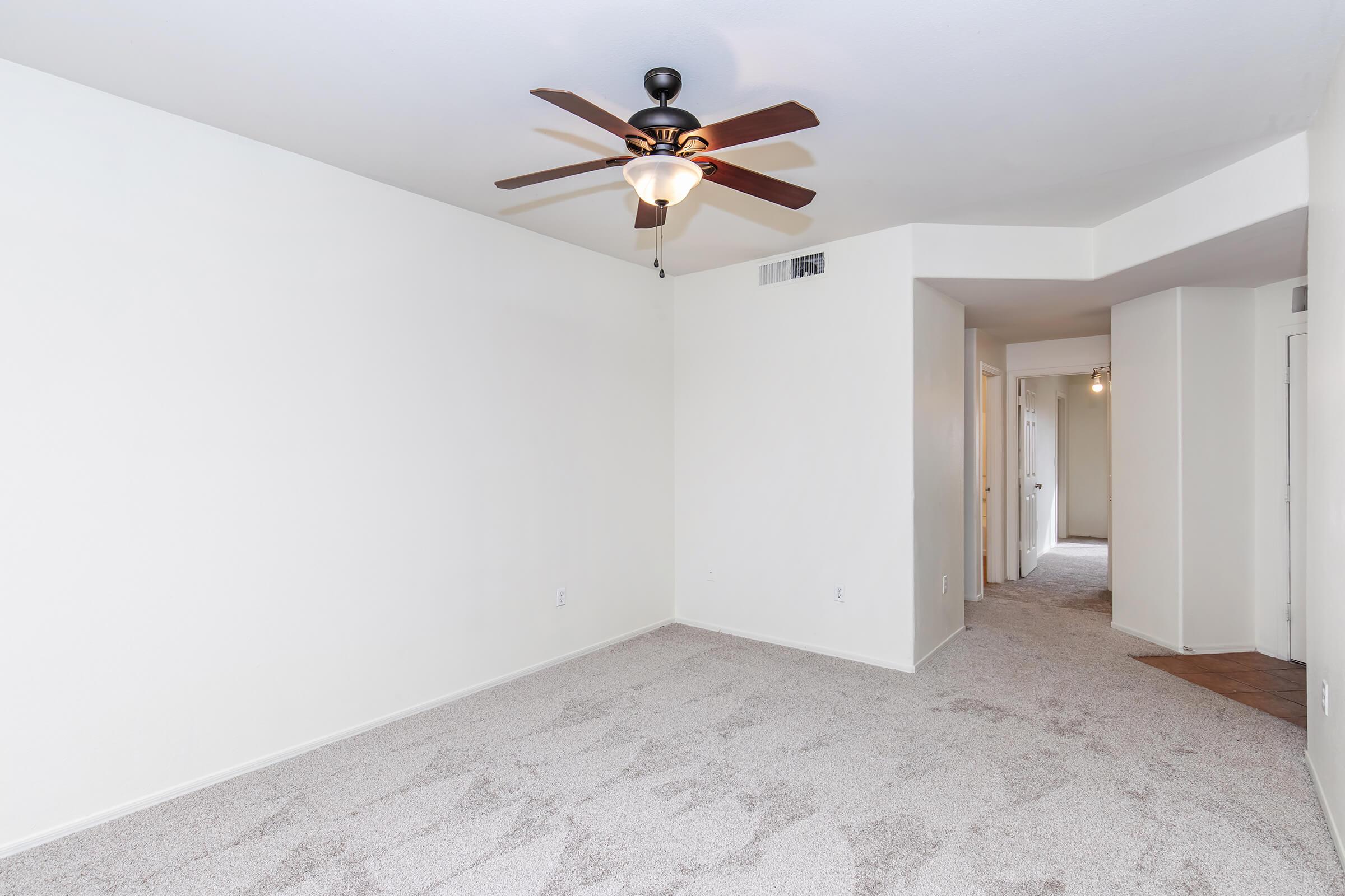 Empty room with light beige walls and plush carpet. A ceiling fan with light fixtures hangs from the ceiling. A hallway leads to additional rooms on the right, visible doorways in the background, and natural light coming from an unseen window. The space appears clean and well-maintained, suitable for various uses.