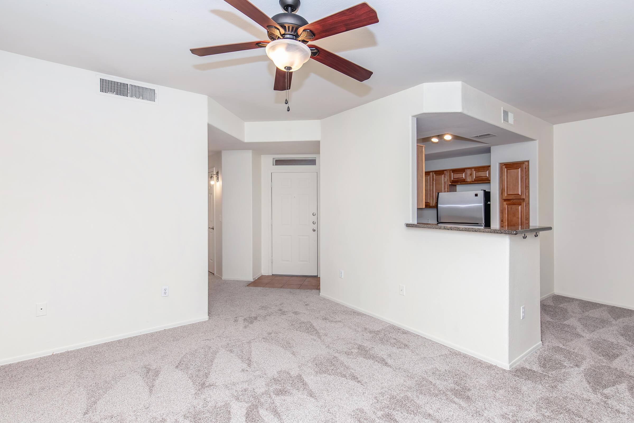 A spacious living area with light-colored walls and plush carpet. A ceiling fan is visible above, and the open layout leads to a kitchen area with wooden cabinets and a stainless steel refrigerator. The entrance door is in the background, showcasing a bright and inviting atmosphere.