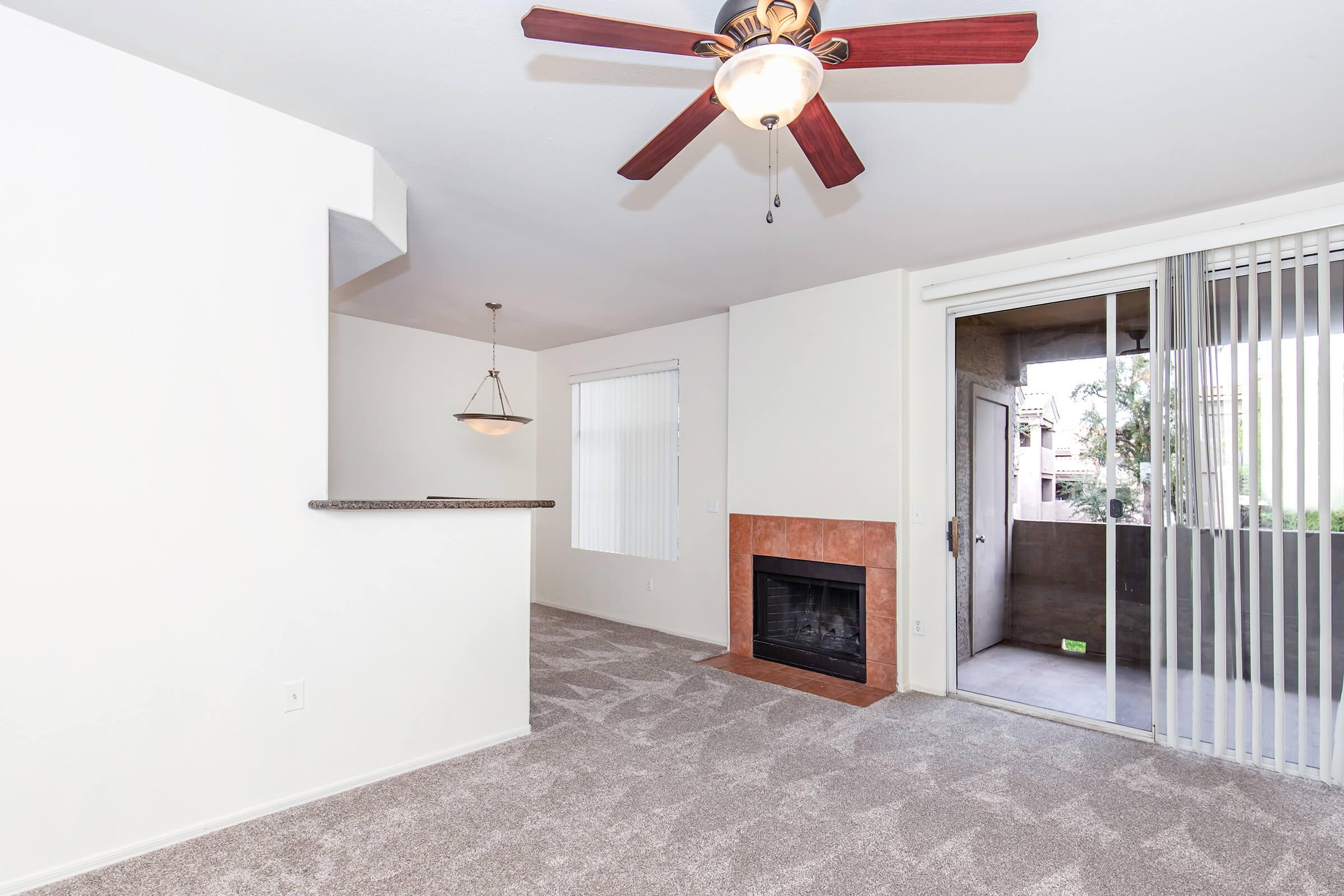 Interior view of a living room featuring light-colored walls, a ceiling fan with wooden blades, and beige carpet. There’s a fireplace with a tiled surround and a sliding glass door leading to a balcony with vertical blinds. A light fixture hangs above a bar counter separating the living area from the kitchen.