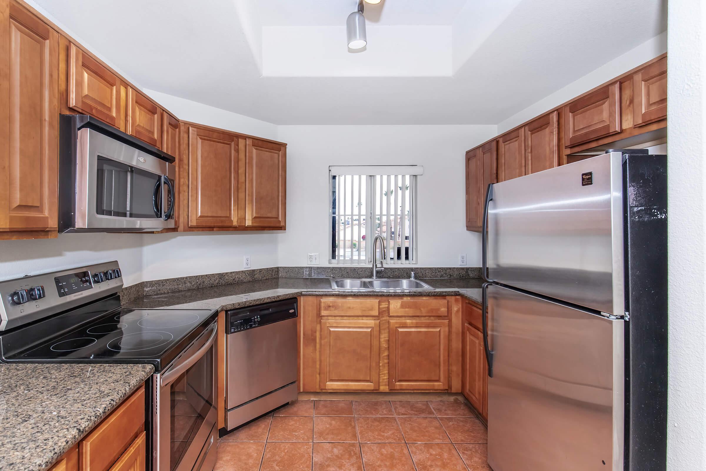 A modern kitchen featuring wooden cabinets, stainless steel appliances, a granite countertop, and tile flooring. The kitchen includes an oven, microwave, dishwasher, and refrigerator. A window with blinds provides natural light.