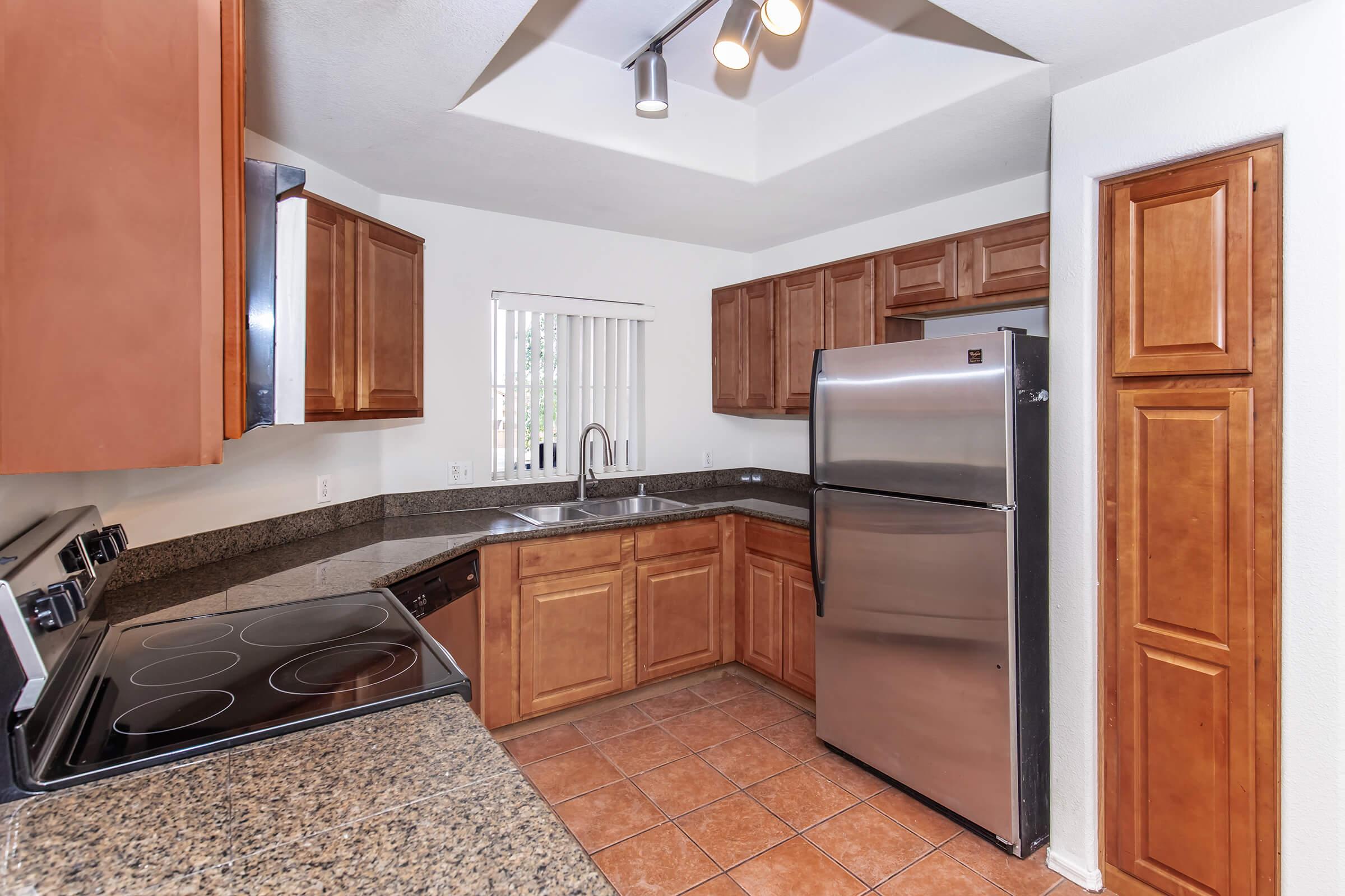 Modern kitchen featuring wooden cabinetry, stainless steel refrigerator, and granite countertops. The space includes a stove and oven, a single basin sink, and tile flooring. Natural light enters through a window with blinds, creating a bright and functional cooking environment.