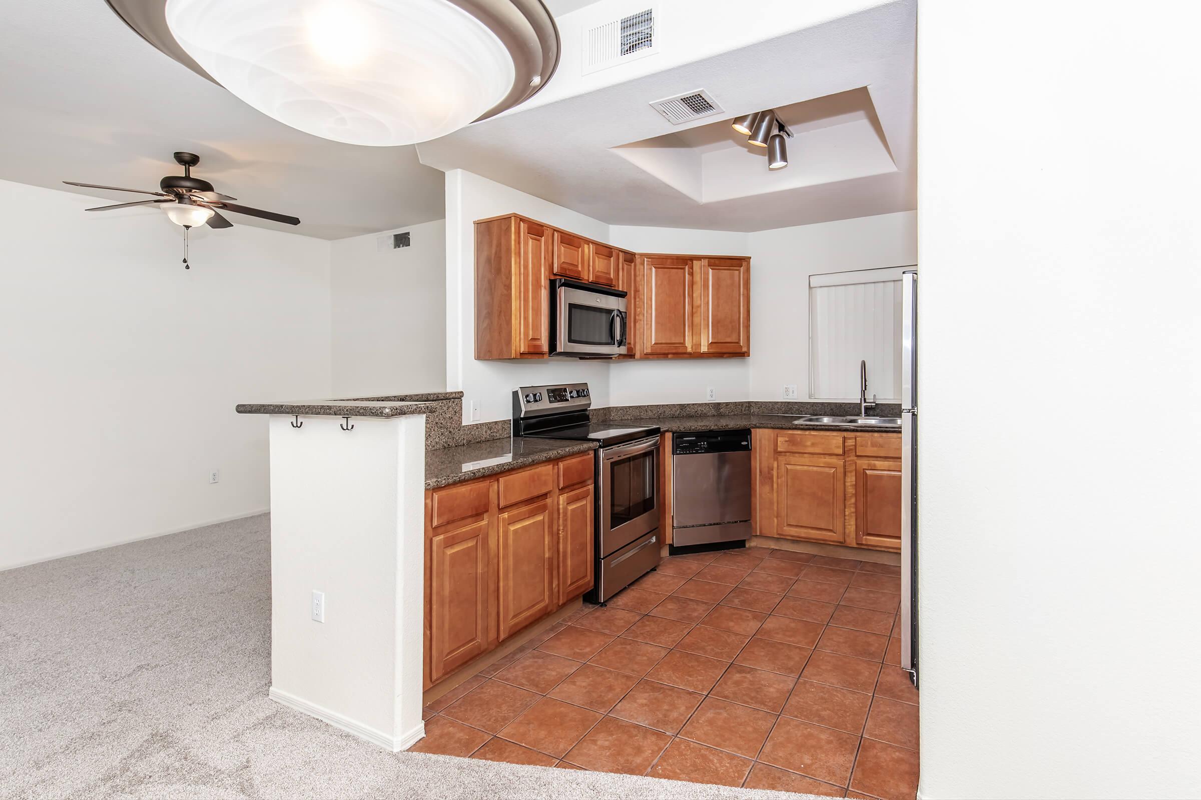 Modern kitchen with wooden cabinetry, stainless steel appliances, and granite countertops. Features include a ceiling fan, a light fixture, and a spacious open layout leading into a living area with carpeted flooring. The kitchen offers an island and an adjacent dining area with a window.