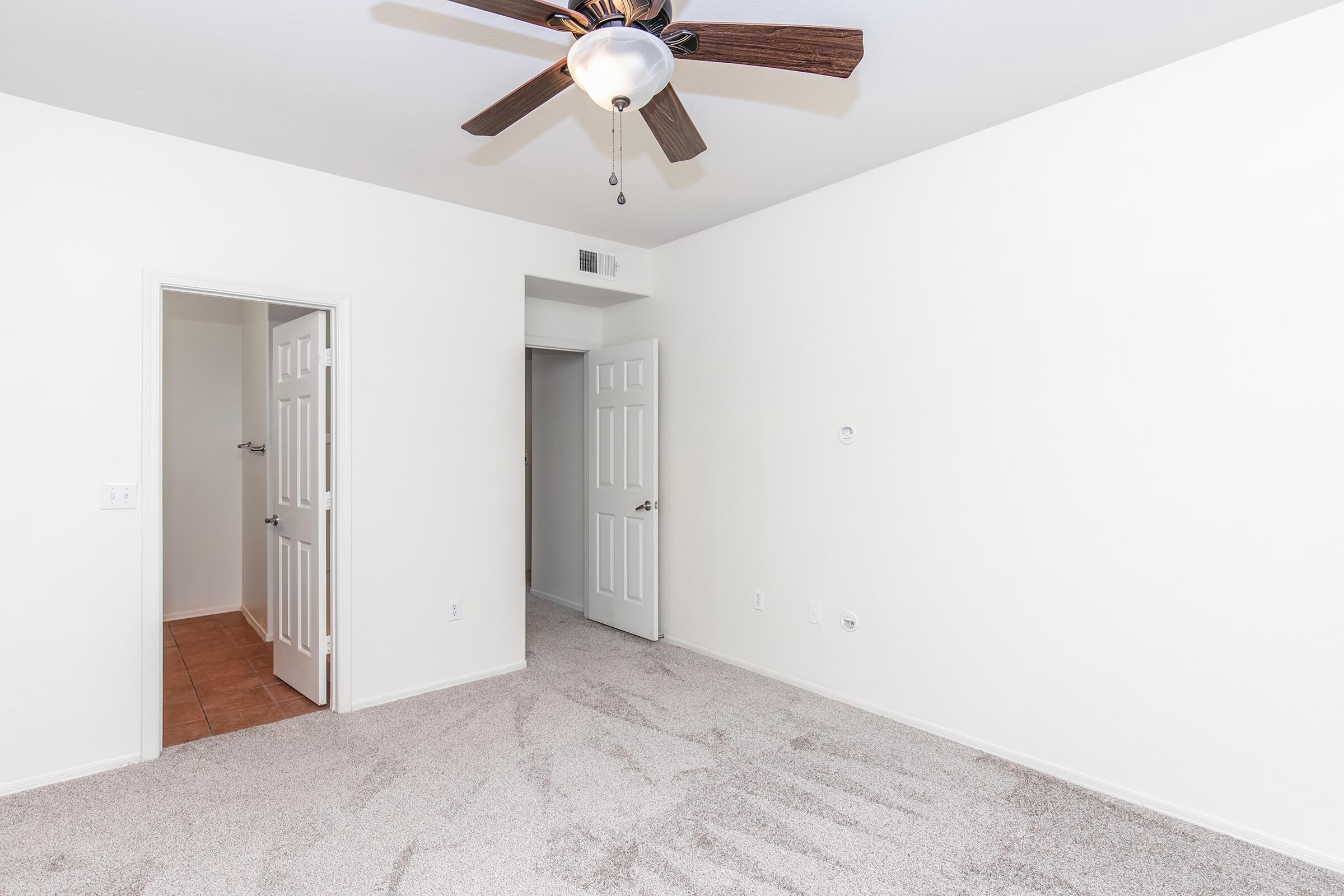Empty room with light-colored walls and beige carpeting. A ceiling fan with wooden blades is installed. Two doorways lead to other spaces, one open and the other partially closed. The room is well-lit, giving a spacious feel with minimal decor.