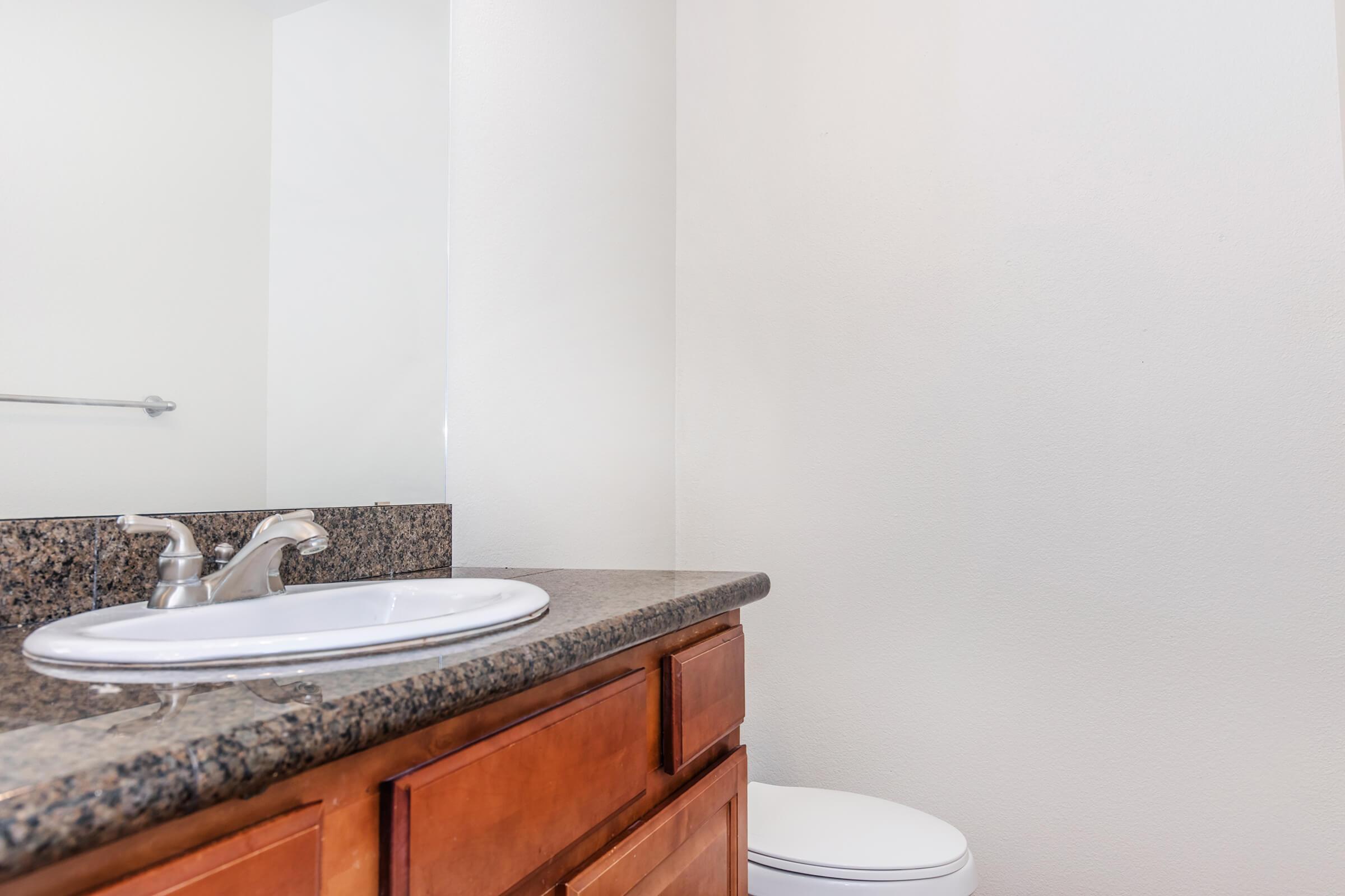 A bathroom with a granite countertop, a white sink, and a faucet. In the background, there is a mirror and a white toilet. The walls are painted in a light color, creating a clean and spacious appearance.