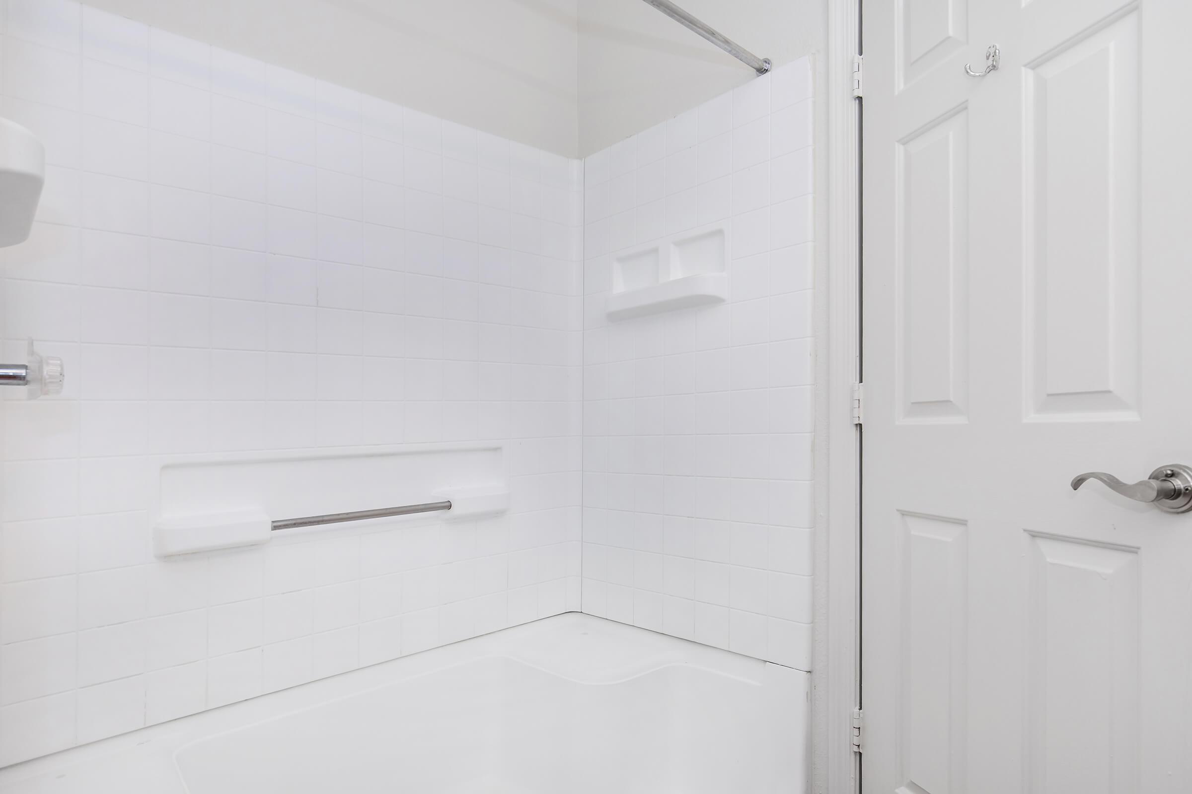 A clean, white tiled bathroom featuring a shower area with a built-in seat and grab bars. The door is closed, and the space is brightly lit, emphasizing the minimalist design with subtle reflections on the tiles.