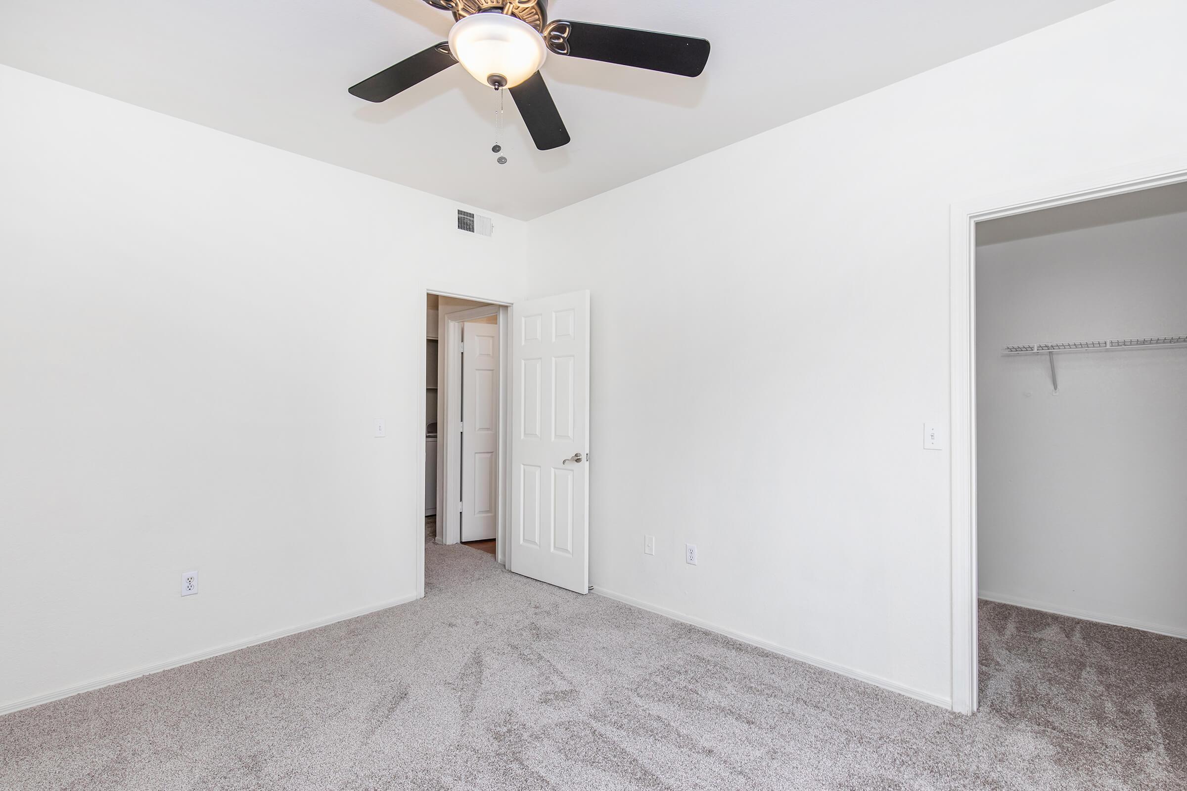 A spacious, empty room with light-colored walls and a ceiling fan. The floor is carpeted in a soft hue. There is a closed door on the left leading to another space and an open closet on the right, showcasing shelving. Natural light brightens the area, creating an inviting atmosphere.
