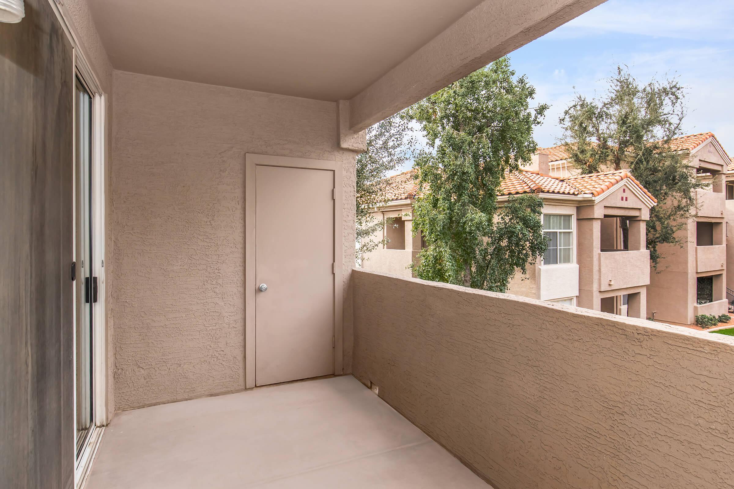 View of a balcony with a beige stucco wall, featuring a closed door on one side. The balcony overlooks a landscaped area with trees and other buildings in the background, showcasing a sunny day.