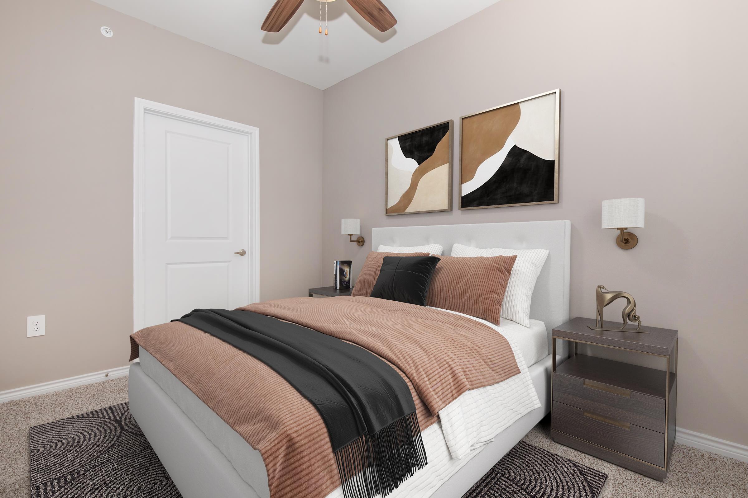 A cozy, elegantly decorated bedroom featuring a queen-sized bed with brown and white bedding, two bedside lamps, and abstract wall art. A small nightstand holds a decorative item. The walls are painted in a soft neutral tone, and there is a ceiling fan above for added comfort.