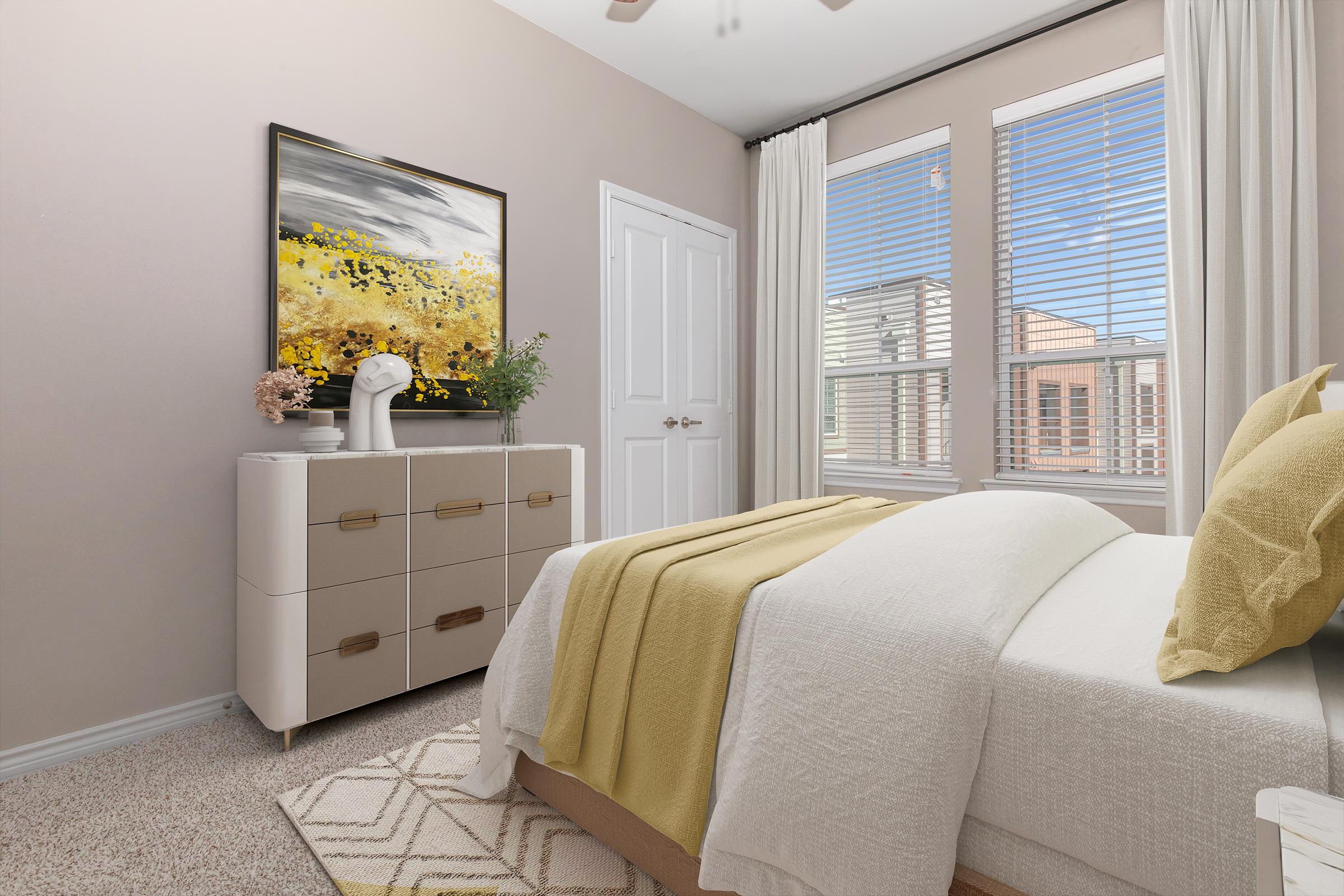 Bright and airy bedroom featuring a neatly made bed with yellow accents, a light-colored comforter, and a decorative throw. A modern dresser with gold handles sits against the wall, and there's a large abstract painting above it. Sunlight streams through window with blinds, creating a warm atmosphere.