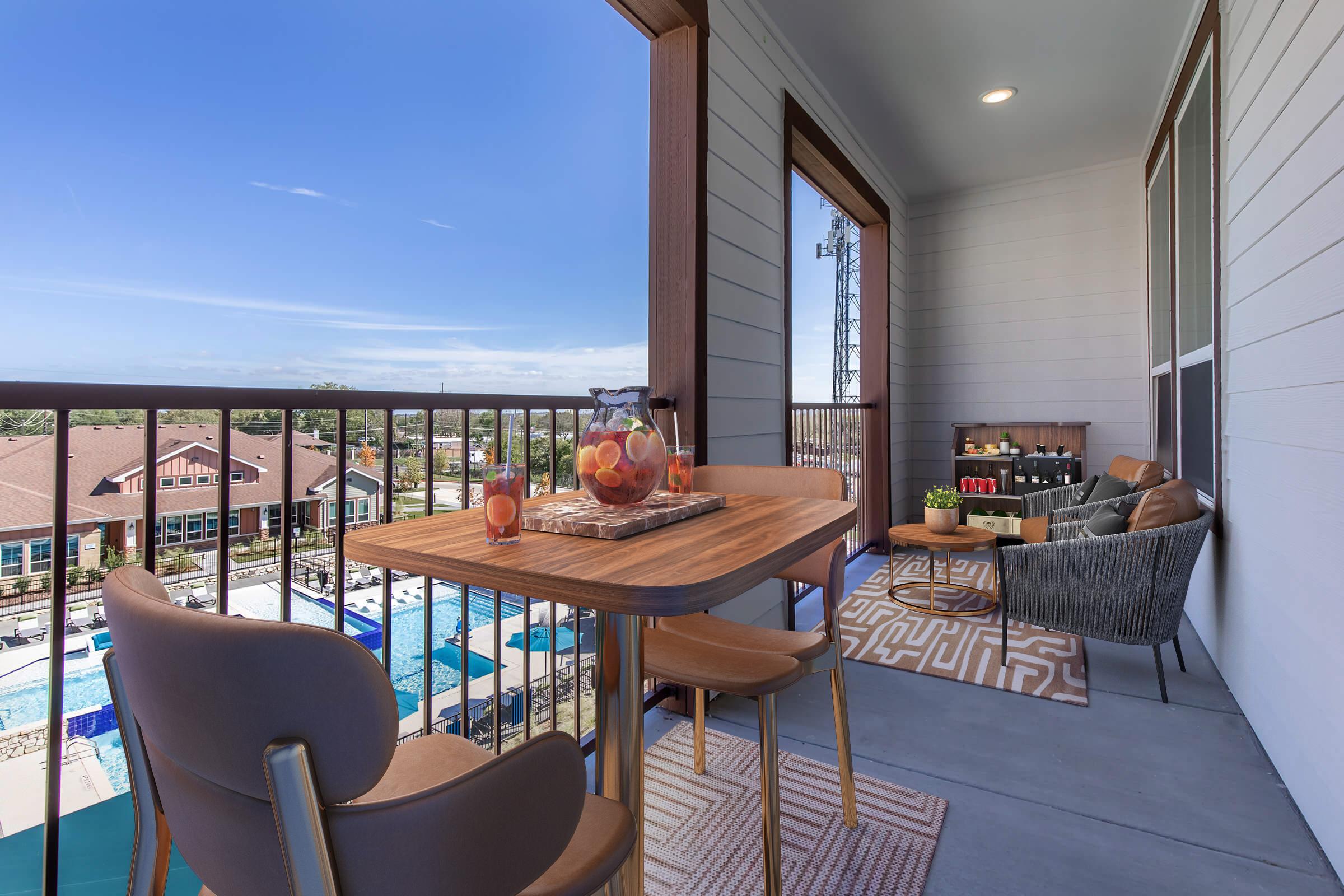 A cozy balcony setting featuring a wooden table with decorative fruits and two stylish chairs. In the background, a view of a pool area and lush landscaping under a clear blue sky. The space is elegantly designed for relaxation and enjoying the outdoors.