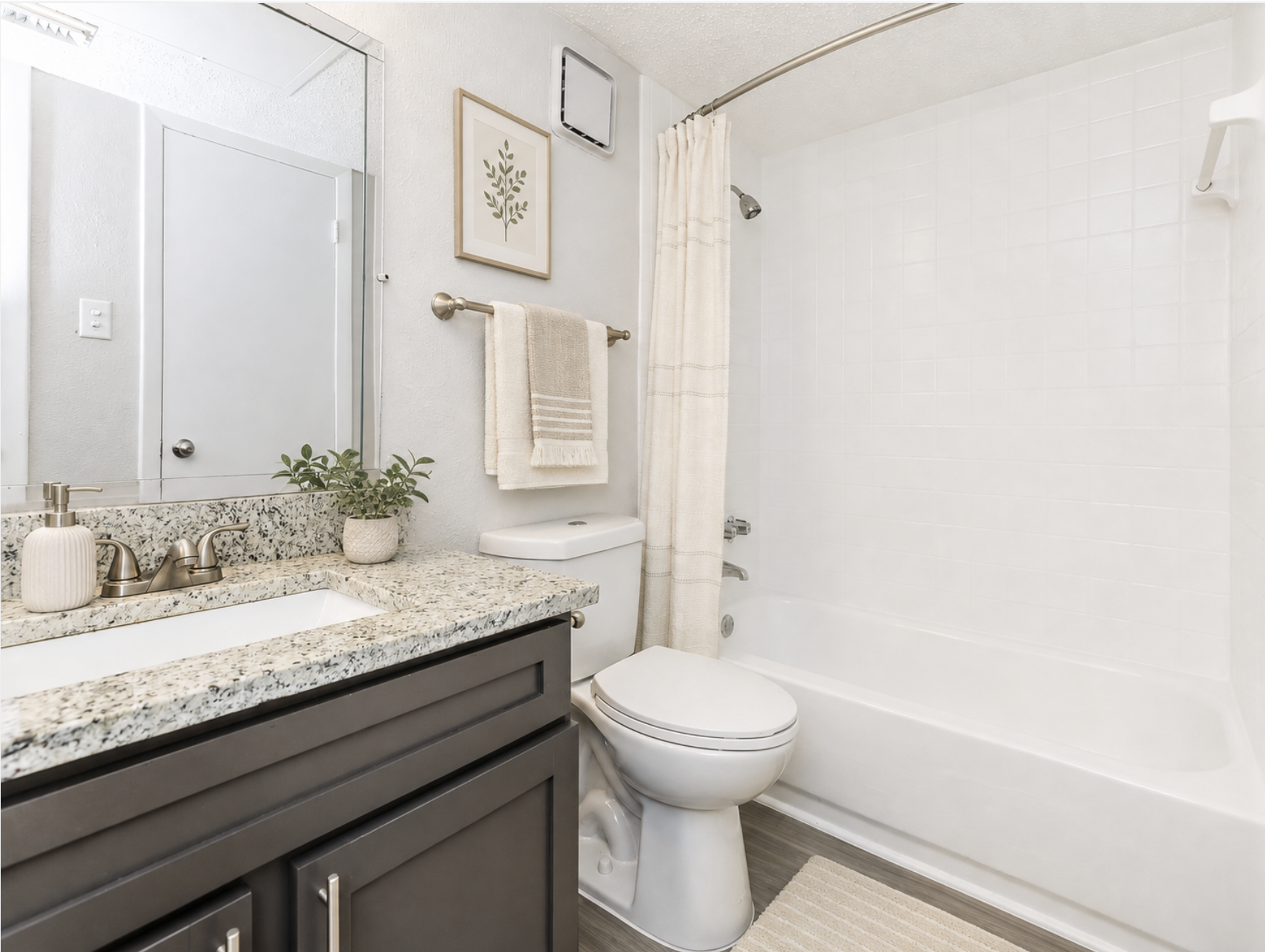 A modern bathroom featuring a rectangular bathtub with a white shower curtain, a toilet, and a granite countertop with a sink. The decor includes a potted plant and a framed artwork on the wall. The walls are painted in a light color, and there is a towel hanging on a bar near the sink.