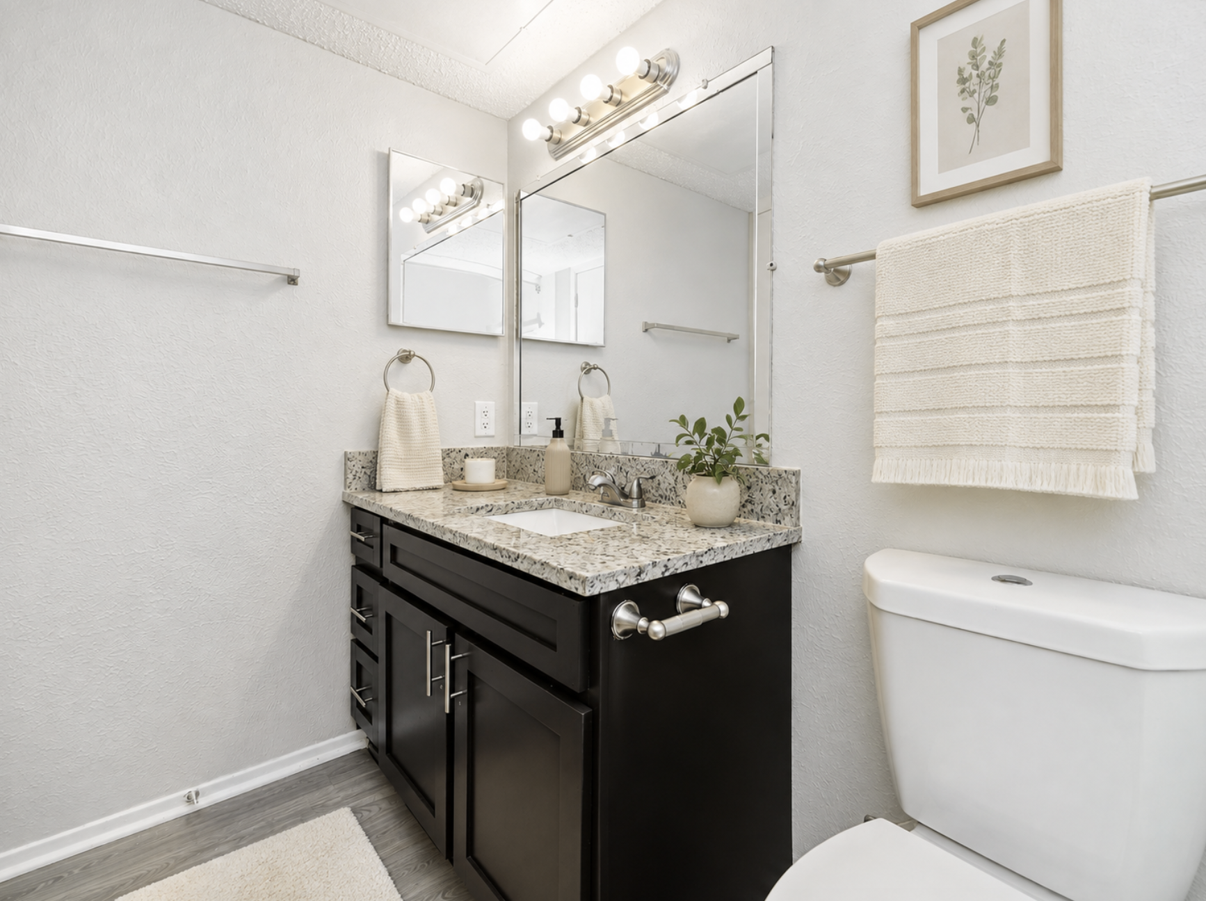 A modern bathroom featuring a dark wood vanity with a granite countertop, a large mirror, and a stylish light fixture above. A small plant and decorative items adorn the countertop. There is a white toilet and a towel rack with a neatly folded towel. The walls are light-colored, creating a bright and airy atmosphere.