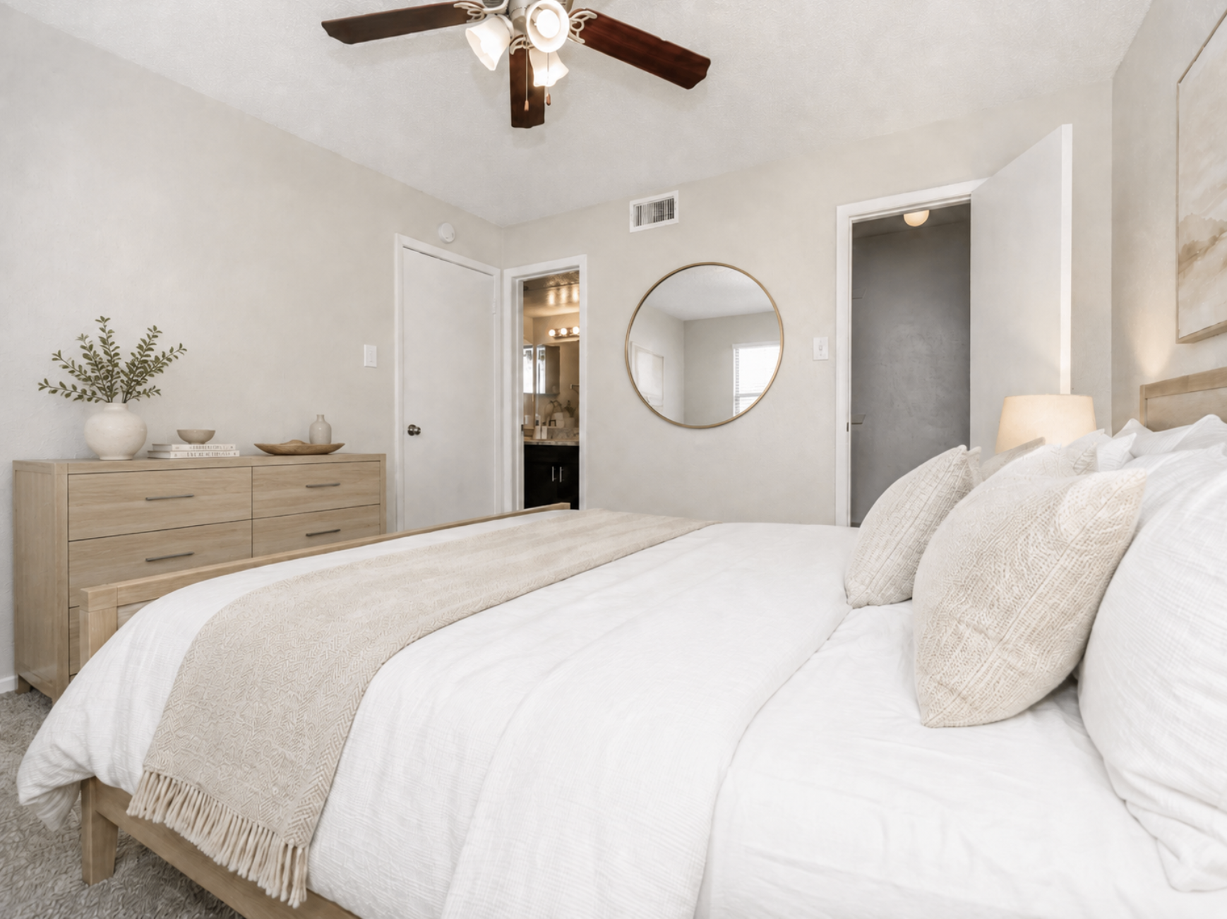 A cozy bedroom featuring a large bed with white linens and decorative pillows, a wooden dresser, and a round mirror on the wall. A ceiling fan adds to the ambiance, while a small plant and decorative items are placed on the dresser. A door leads to a bathroom, visible in the background.