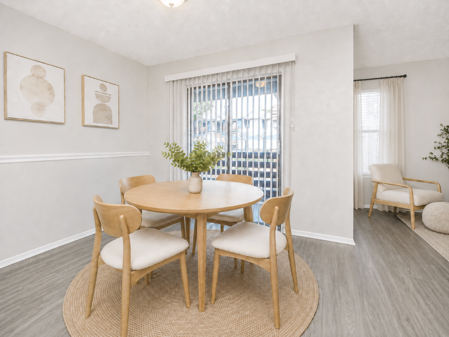 A cozy dining area featuring a round wooden table surrounded by four light-colored chairs. A vase with greenery sits on the table. The walls are painted in a soft tone, and there are two abstract art pieces. Large sliding glass doors allow natural light, leading to a balcony. A beige rug completes the decor.