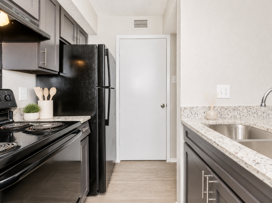 A modern kitchen featuring dark cabinetry, a black stove, and refrigerator. The countertops are made of light-colored granite, and the floor is a light wood laminate. A door leads to another area, and there are kitchen utensils on display. The space is well-lit and neatly arranged.