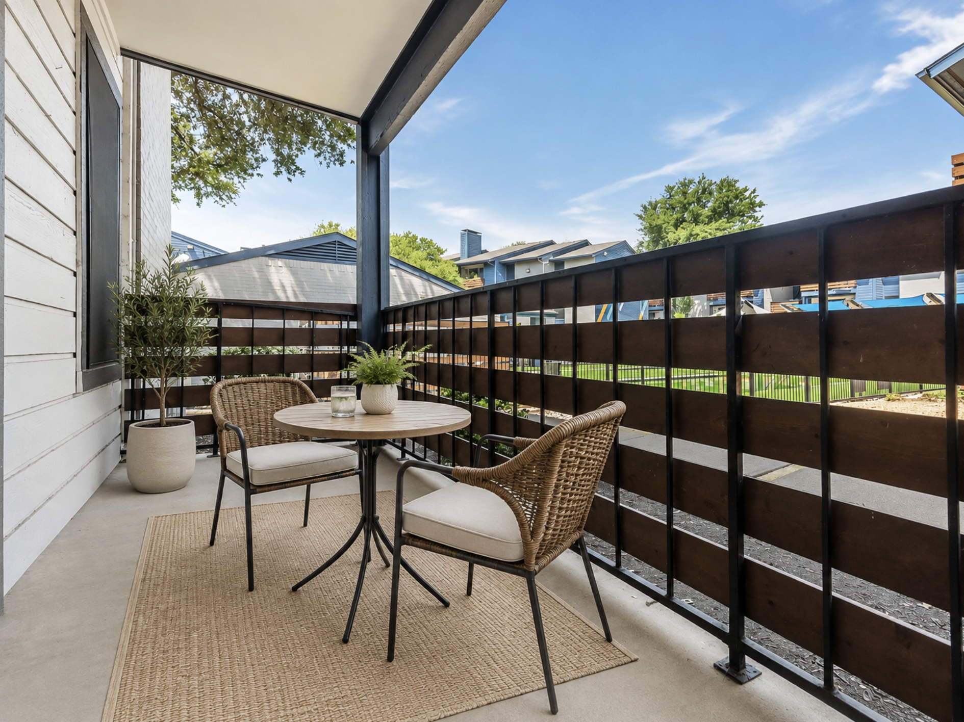 A cozy balcony area featuring a small round table with two chairs. The floor is covered with a neutral-colored rug, and there's a potted plant on the table. Wooden privacy slats line the balcony, with greenery and buildings visible in the background under a clear blue sky.