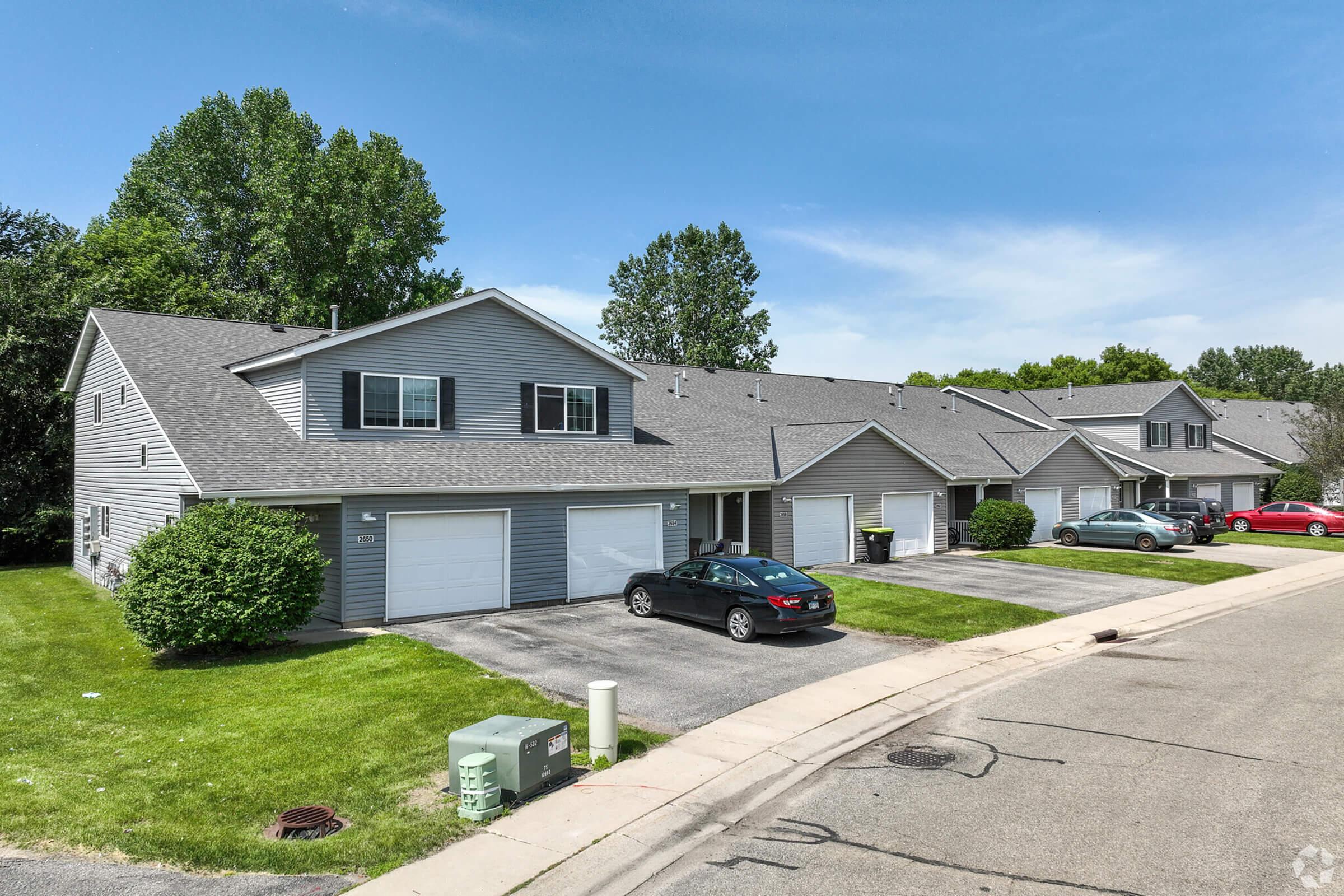 A row of gray residential buildings with multiple garages, surrounded by green lawns and trees. The street curves in the foreground, and several parked cars are visible in front of the homes. The sky is clear with a few clouds.