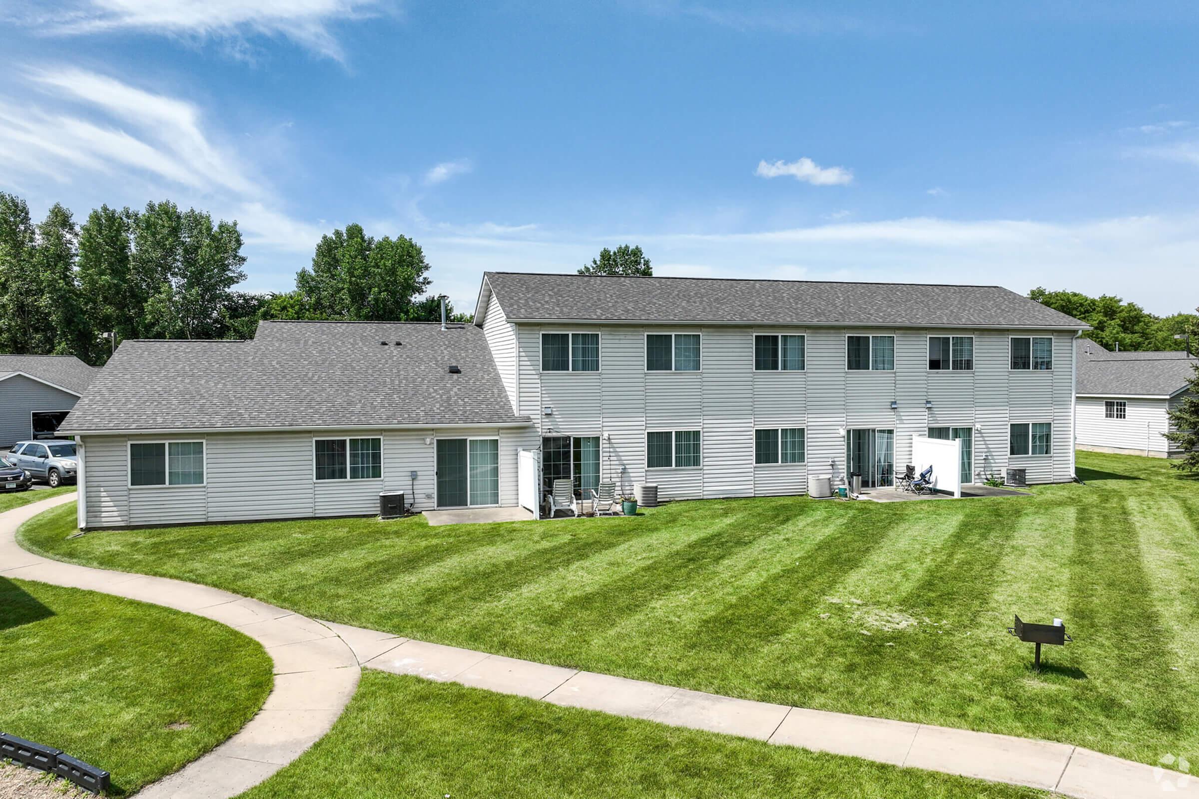 A view of a two-story residential building with multiple units, set in a grassy area. The building features a light-colored exterior with several windows and a porch. Well-maintained landscaping surrounds the building, and there is a paved pathway leading through the lawn. Blue sky and trees are visible in the background.
