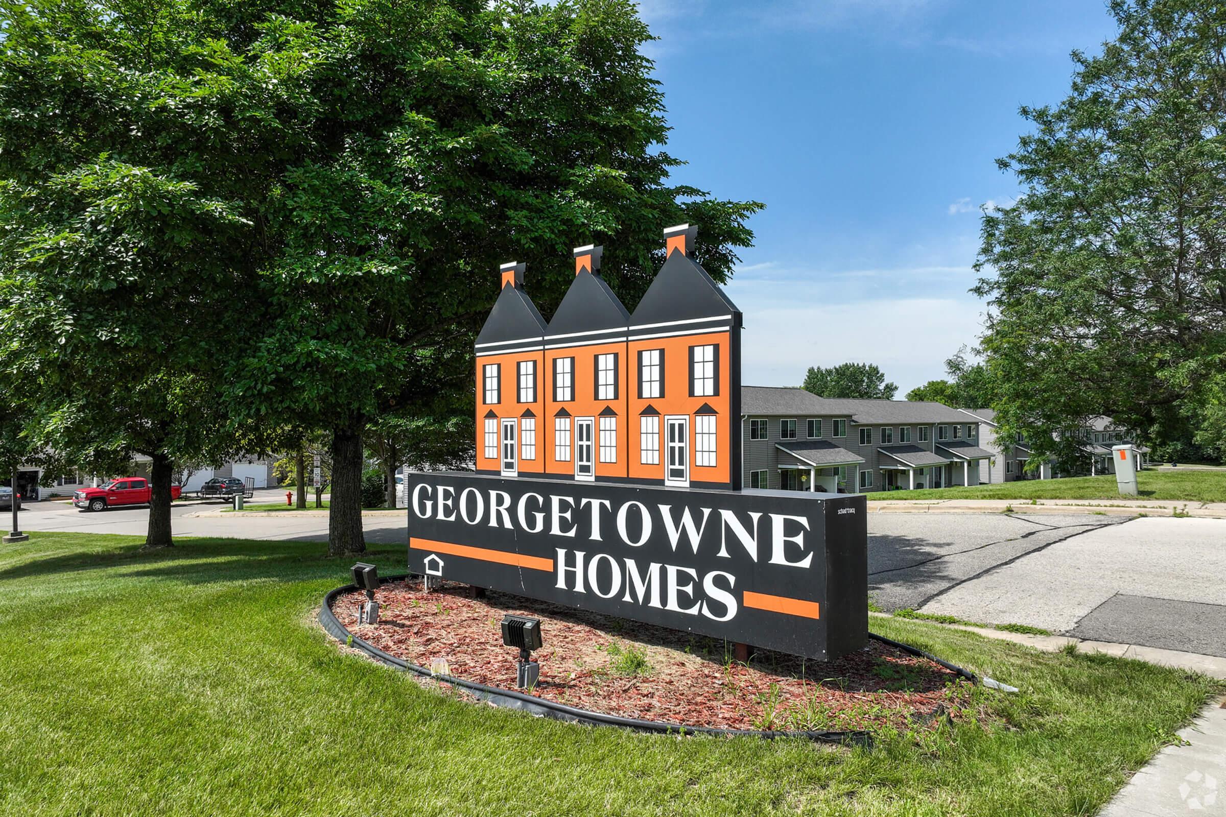 Sign for Georgetowne Homes featuring three orange townhouses with black roofs. The sign is situated in a green landscape with trees and a clear blue sky in the background. Residential buildings can be seen in the distance.