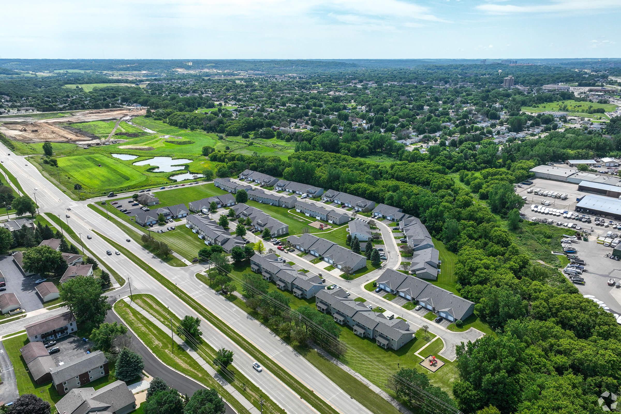 Aerial view of a suburban neighborhood featuring residential buildings arranged in a curved layout. Surrounding green fields and trees, with a pond visible in the distance. A road runs alongside the neighborhood, and industrial or commercial structures can be seen on the right side of the image.
