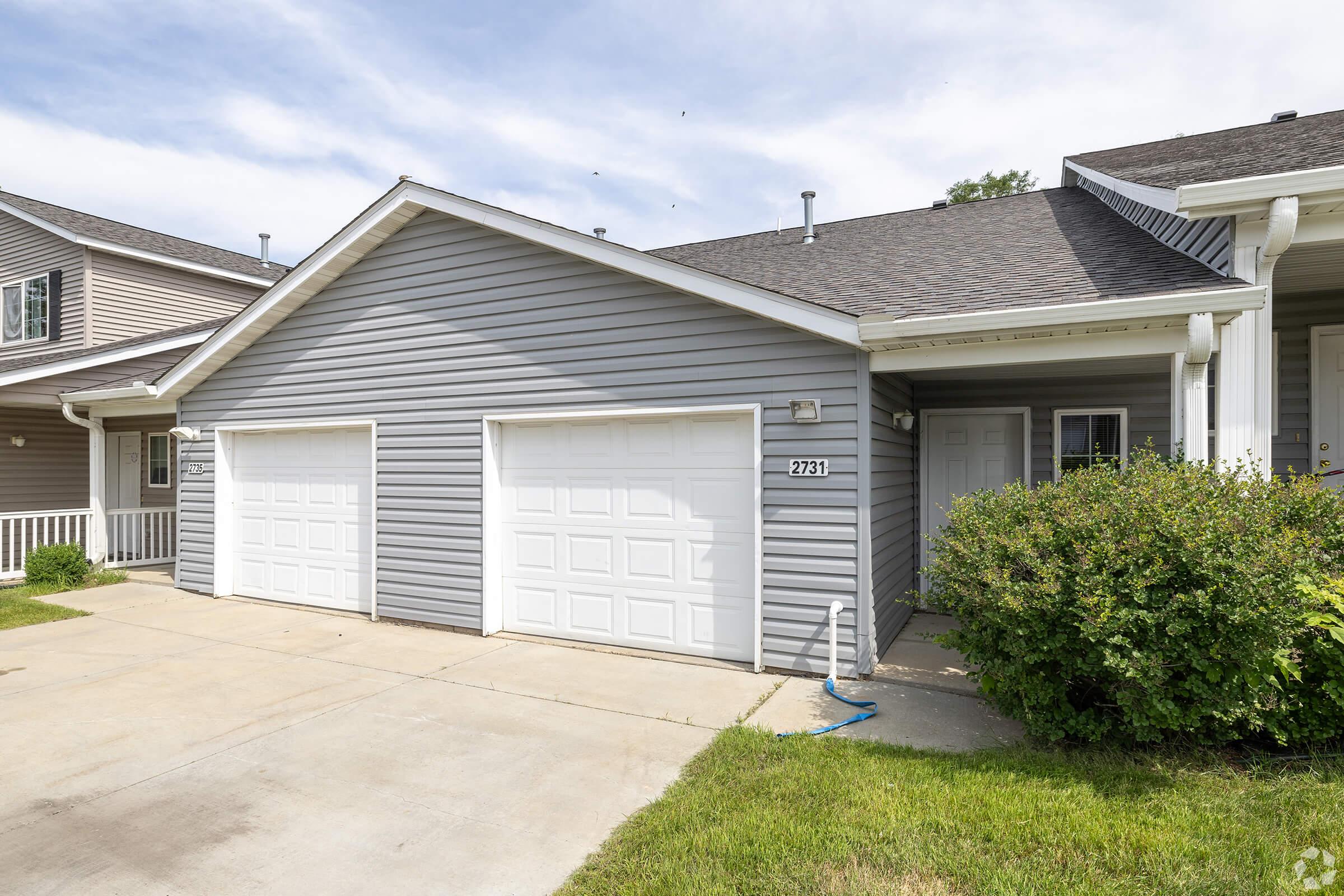 A residential house featuring gray siding, a double garage with white doors, and a walkway. The front yard has neatly trimmed bushes and grass, with a partially cloudy sky in the background. The house number "2731" is visible near the door.