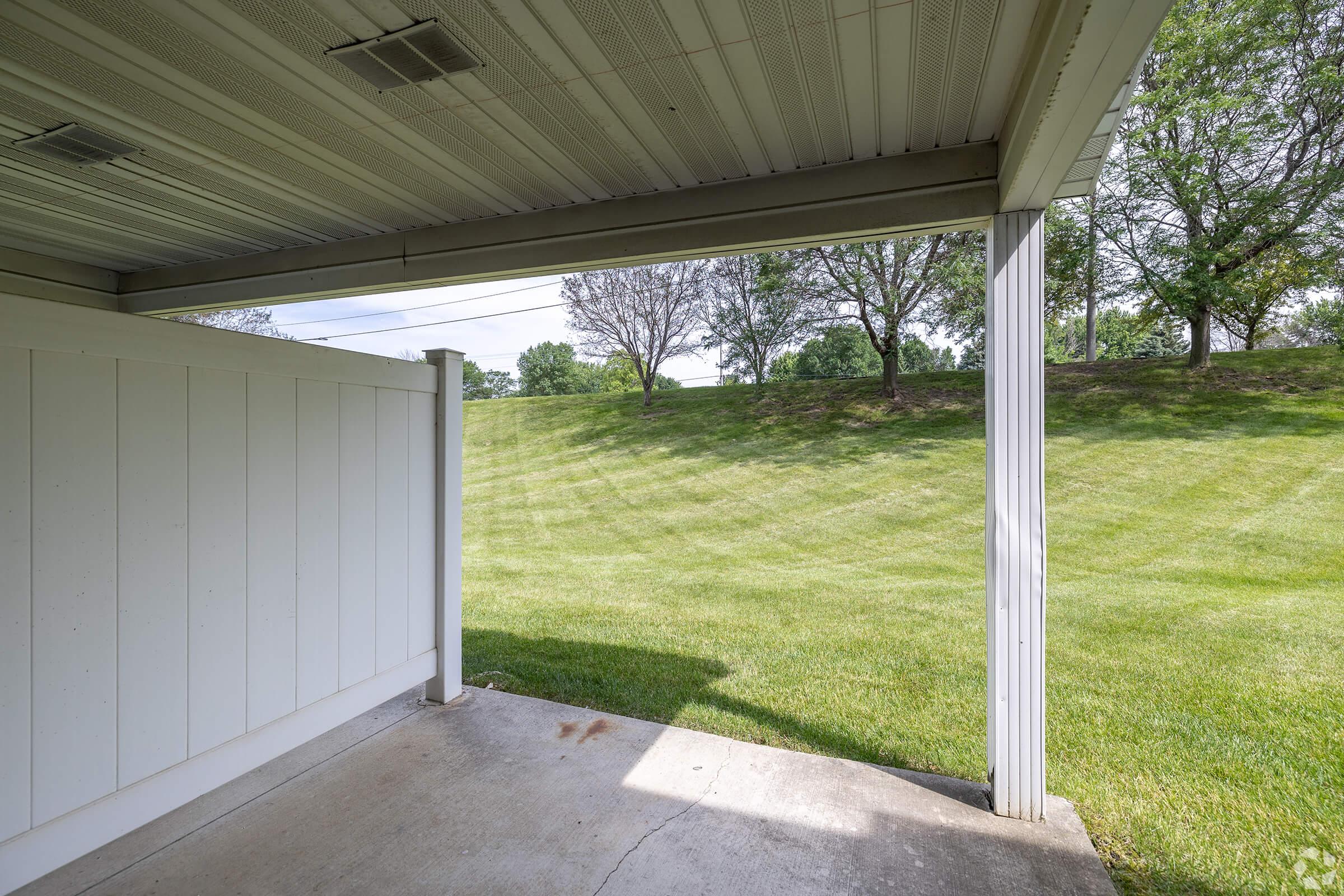 View from a covered patio or porch, showcasing a grassy lawn and trees in the background. The floor is concrete, and there is a white wooden wall to one side. The scene is bright and welcoming, with a clear sky above and well-maintained landscaping.