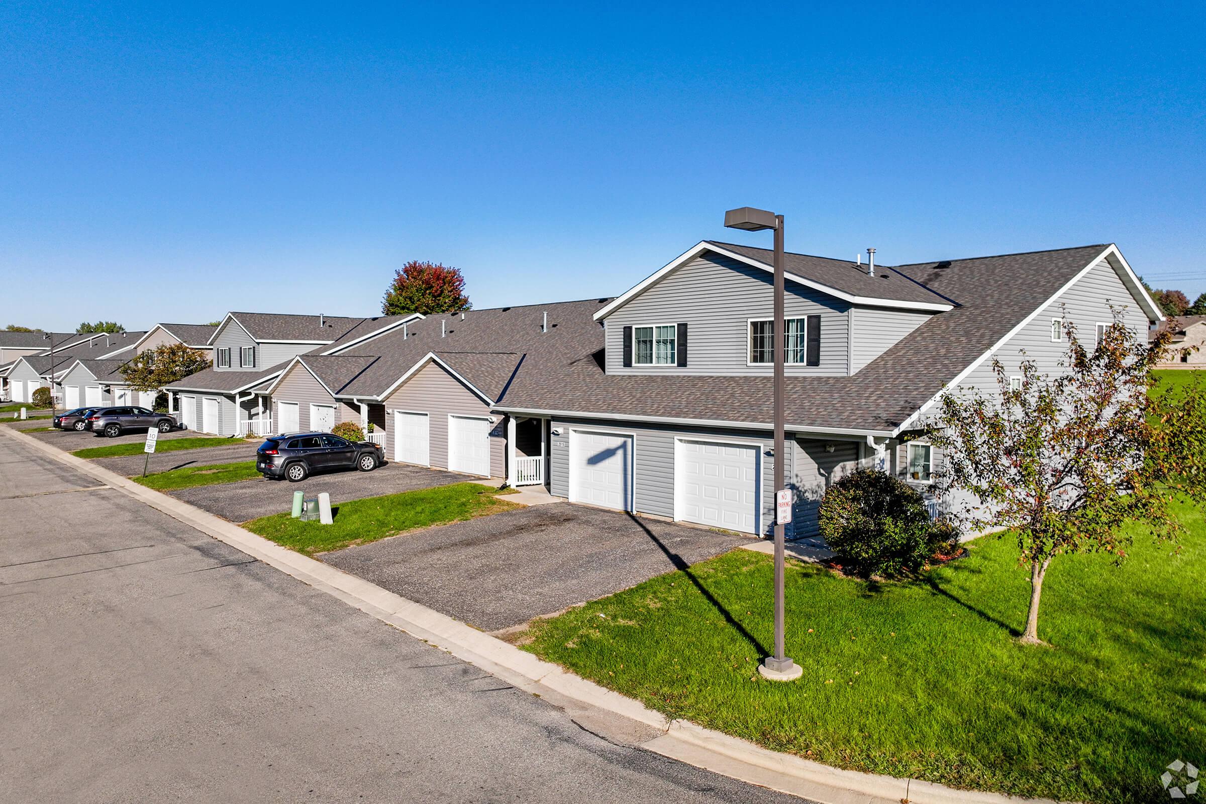 Aerial view of a suburban residential area featuring several attached townhouses with gray siding and pitched roofs. Each unit has a driveway with parked cars, and there are green lawns and trees lining the street under a clear blue sky.