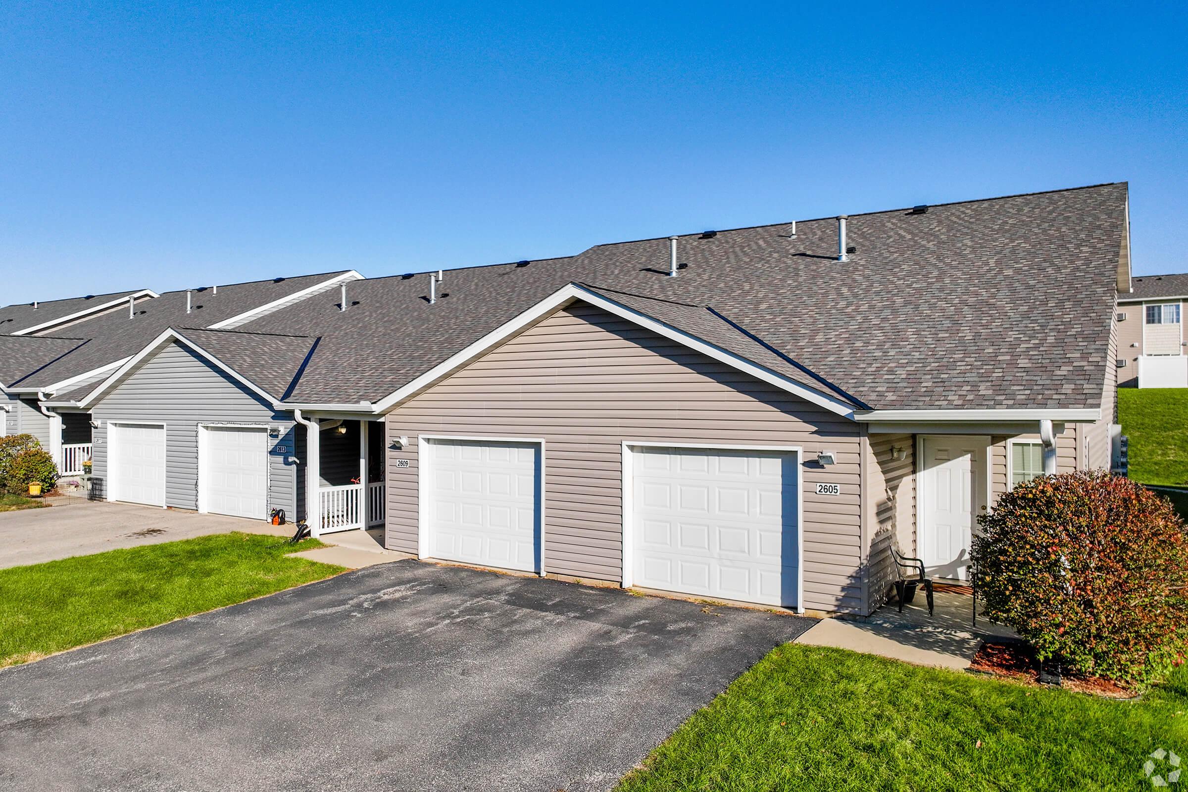 A row of single-story townhouses with gray siding and white garage doors. The houses have sloped roofs and a well-maintained lawn. One home features a small porch with a chair, and the clear blue sky provides a bright backdrop.