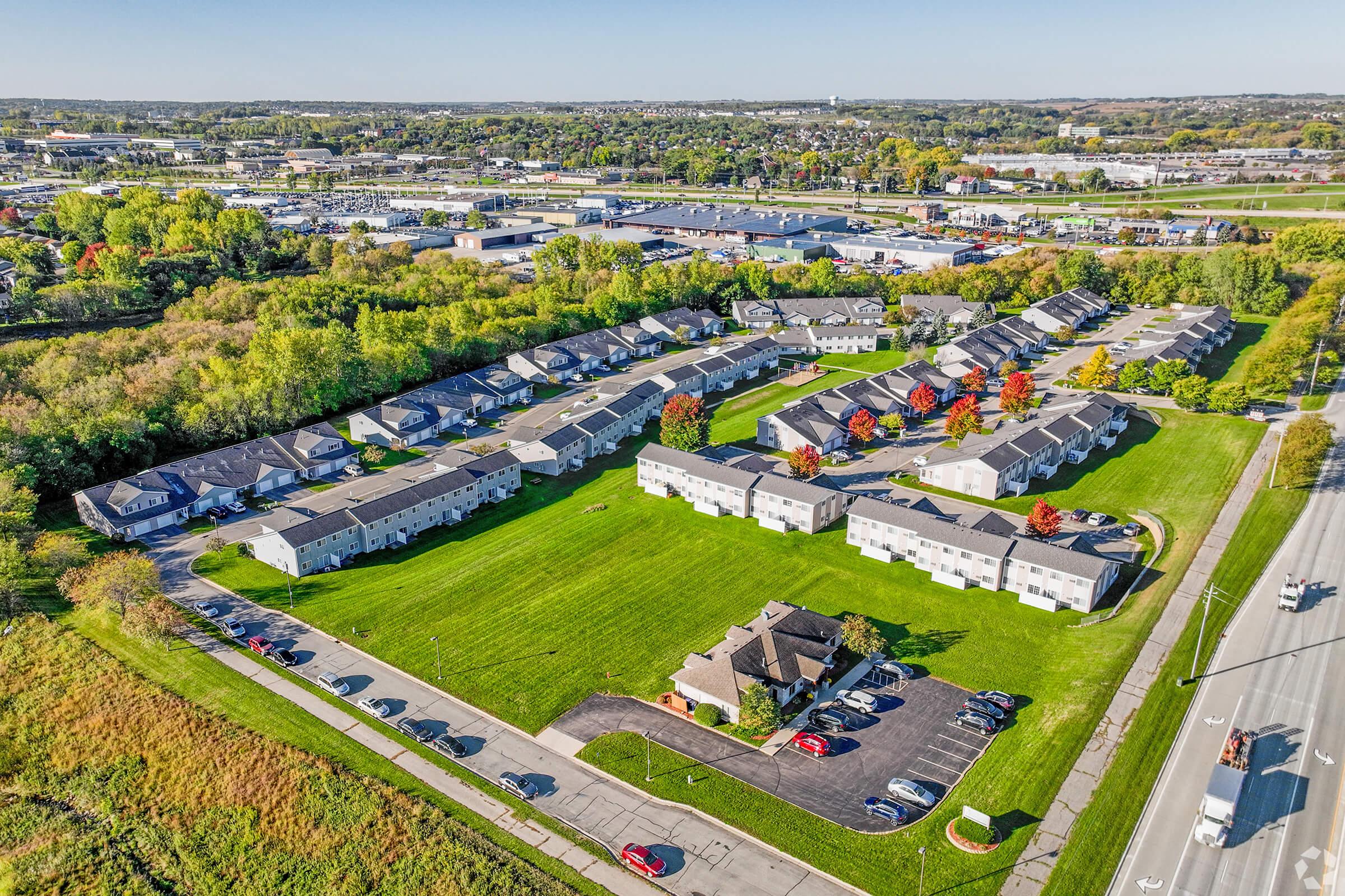 Aerial view of a residential complex featuring multiple buildings arranged in a circular layout surrounded by green lawns and trees. Nearby streets and a parking area are visible, along with industrial buildings in the background. The scene showcases autumn colors in some of the trees.