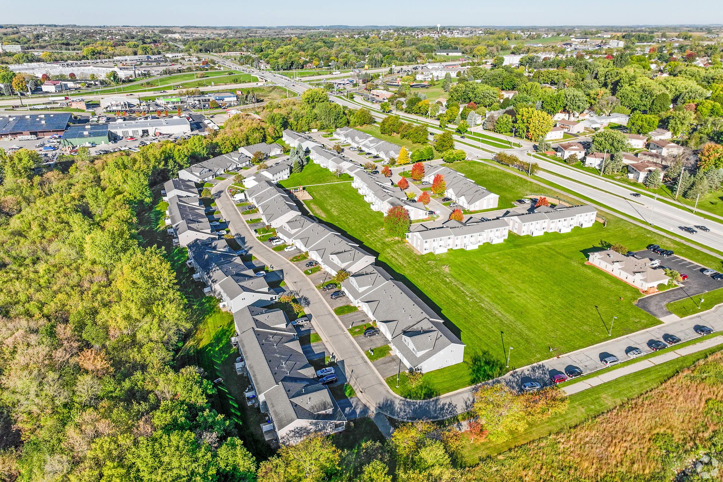 Aerial view of a residential neighborhood featuring a series of low-rise apartment buildings arranged around green lawns and trees. Nearby, a highway and commercial buildings can be seen in the distance, set against a backdrop of clear blue skies and rolling hills.