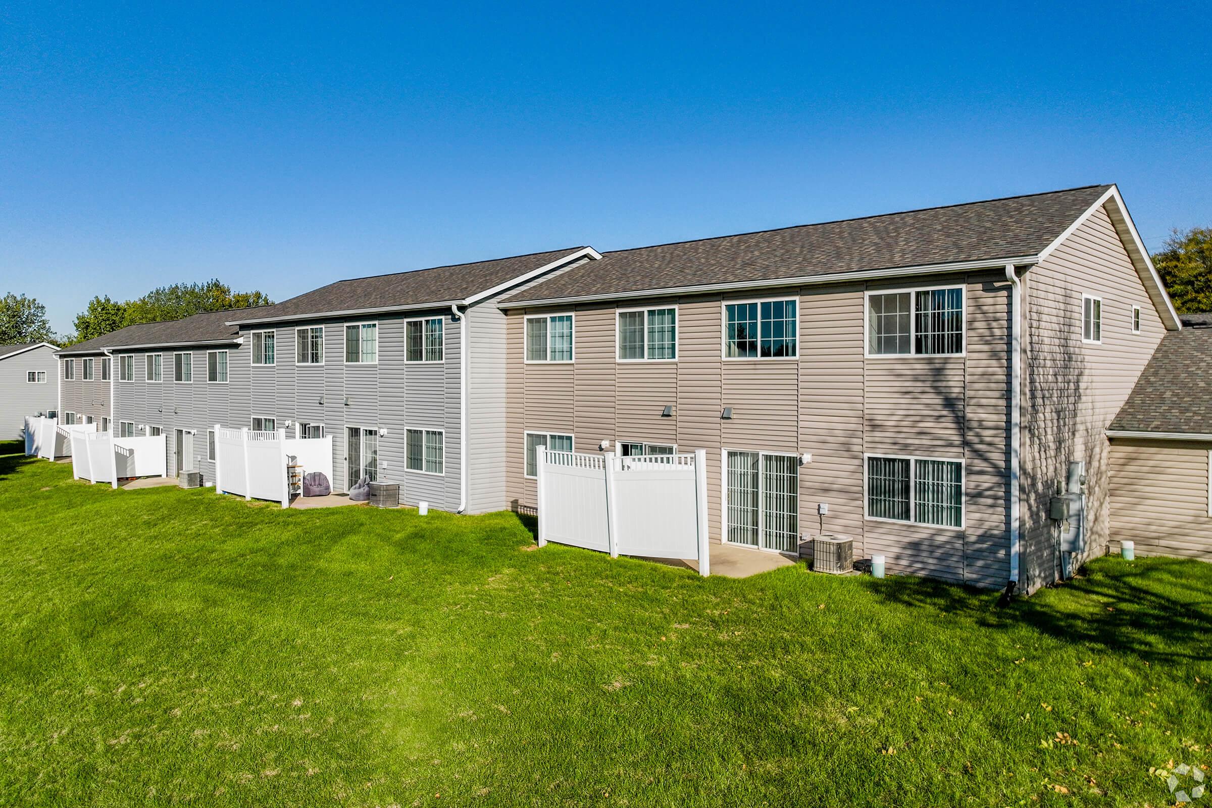 Row of multi-unit residential buildings with a grassy area in the foreground. The buildings feature a mix of light and dark siding, and each unit has a small patio enclosed by white fences. Clear blue sky fills the background, creating a bright and inviting atmosphere.