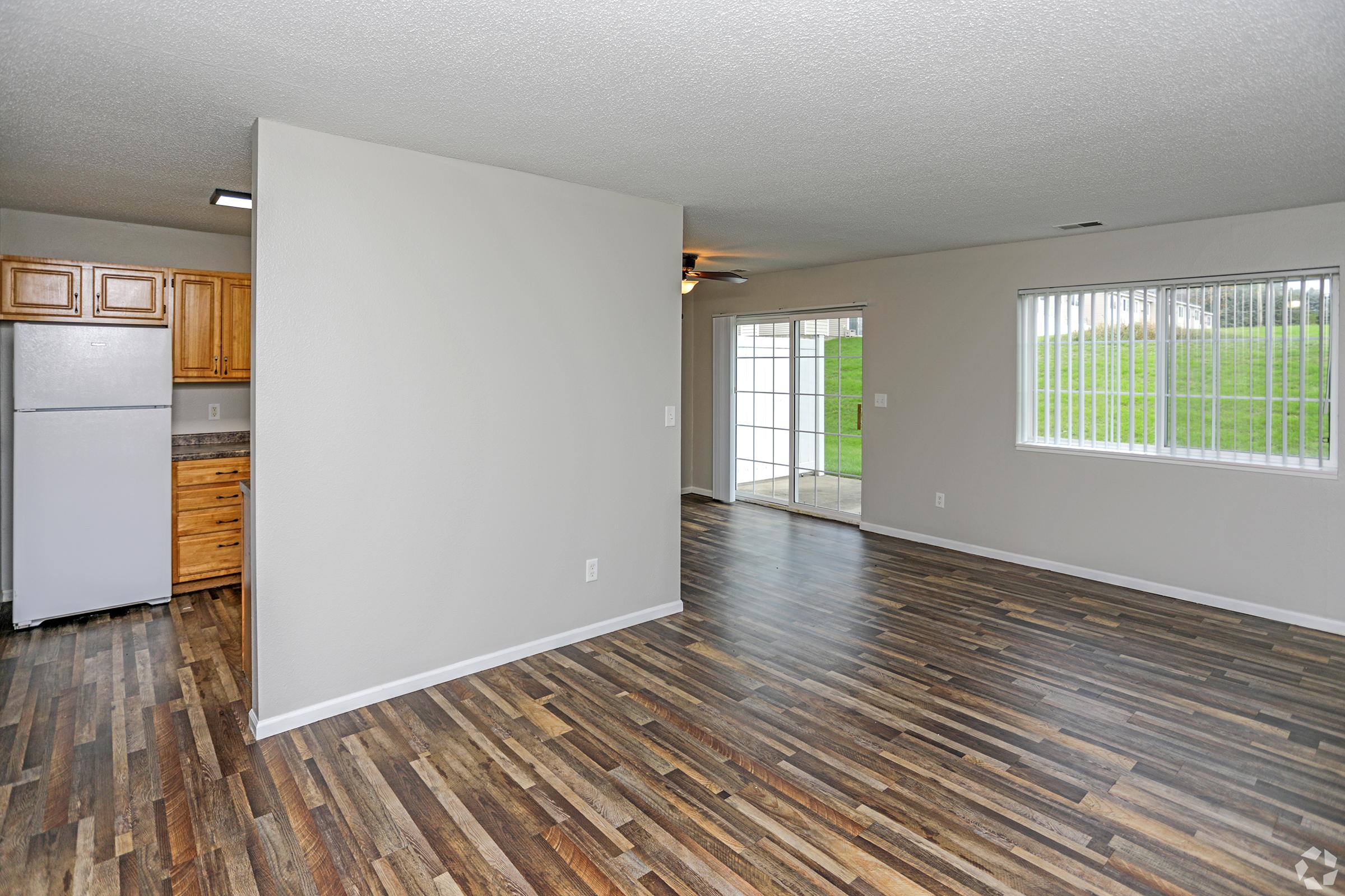 A spacious, modern living area featuring hardwood-style flooring, light-colored walls, a kitchen with wooden cabinets, and a large window letting in natural light. The room is empty, showcasing the potential for various furniture arrangements. A sliding door opens to an outdoor space.